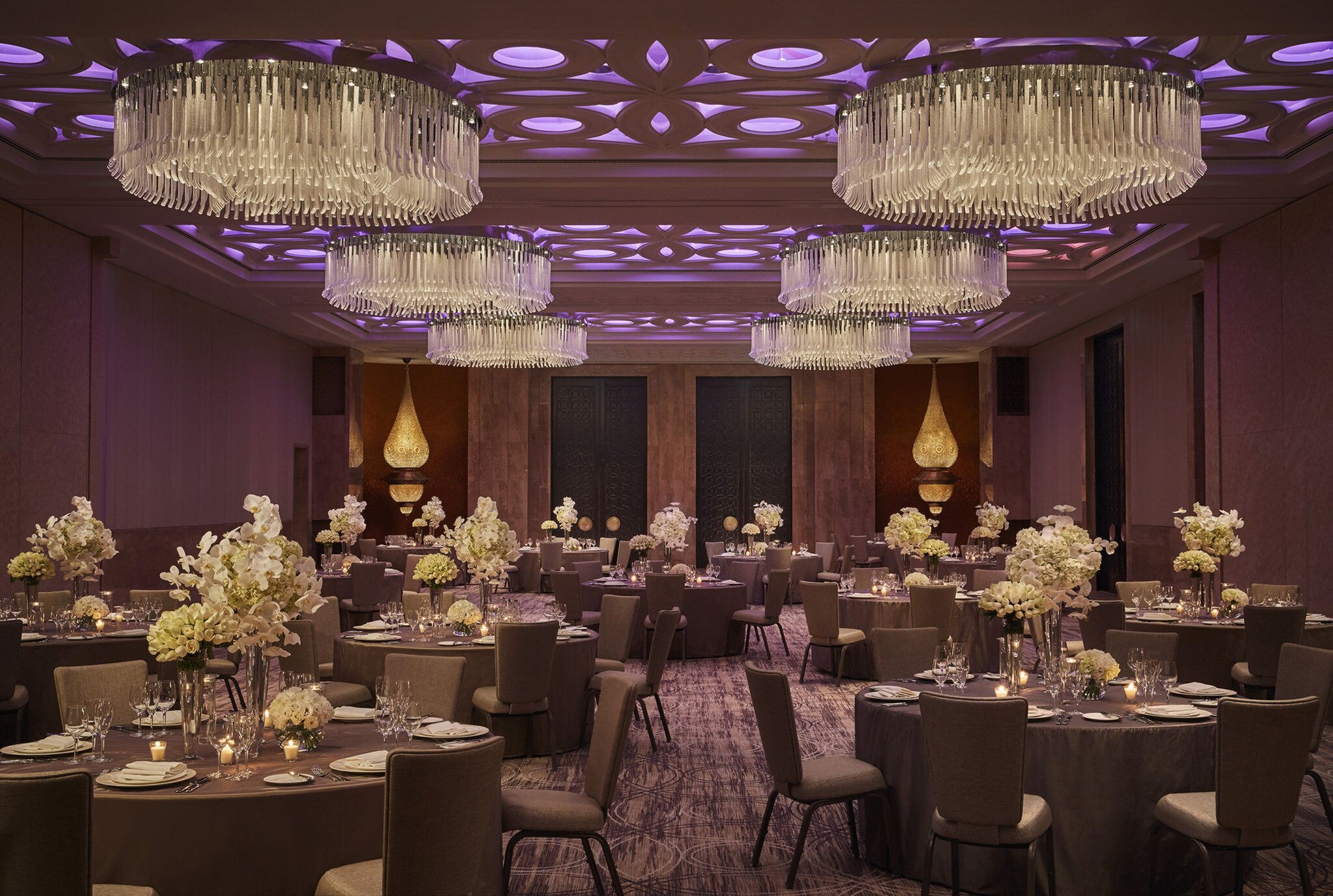 Une salle de banquet dotée de nombreuses tables rondes, chacune agrémentée de fleurs blanches et de bougies, et de grands lustres suspendus au plafond.