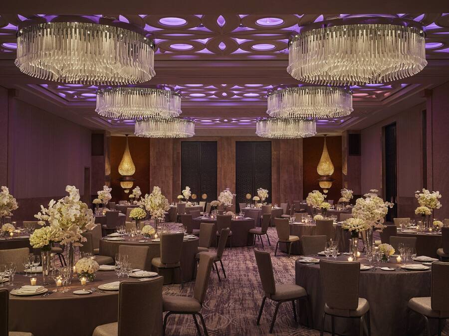 Um salão de banquetes com várias mesas redondas, cada uma decorada com flores brancas e velas, e grandes lustres pendurados no teto.