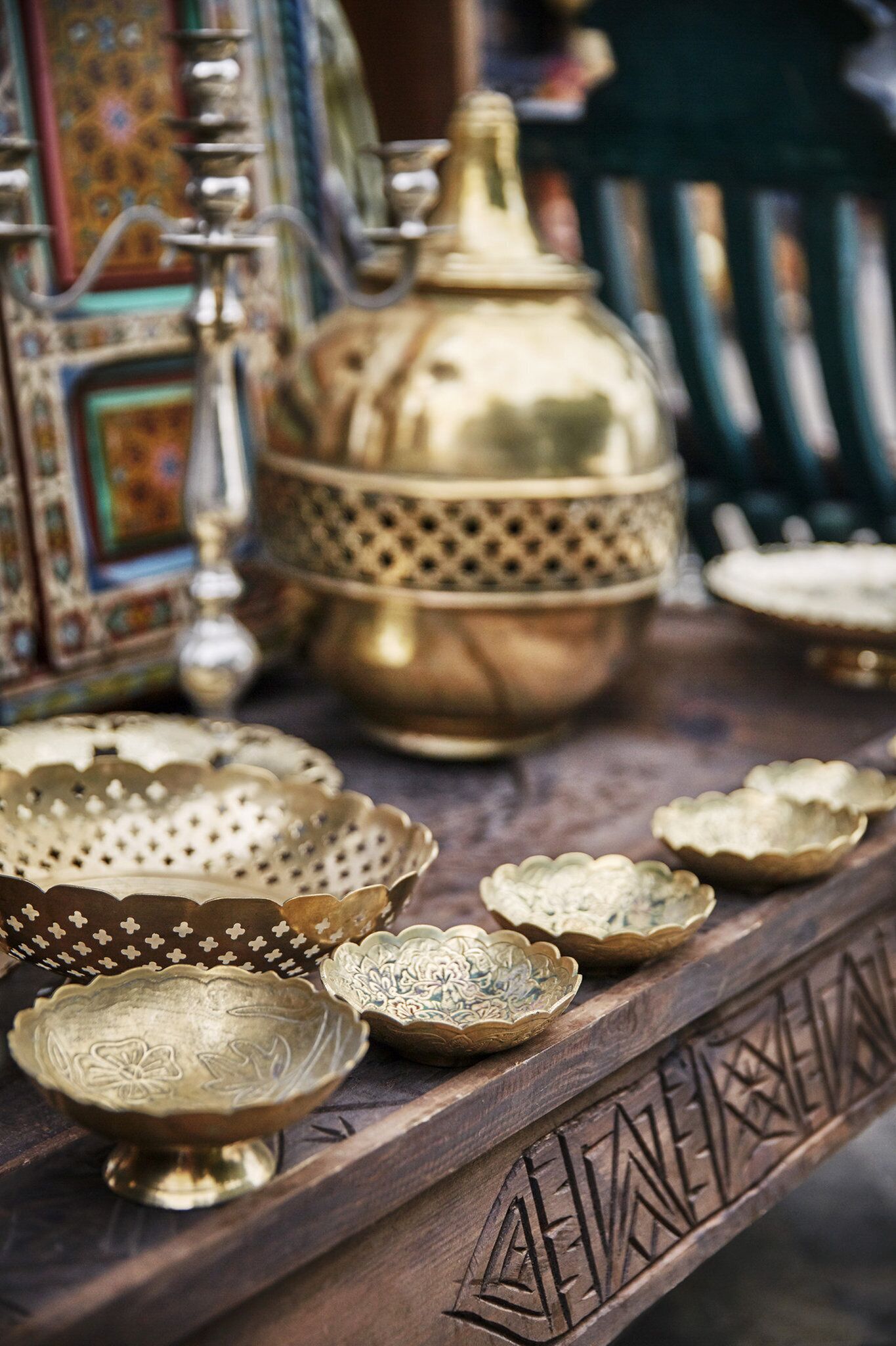 A variety of brass and ceramic bowls and dishes with intricate designs, displayed on a wooden surface with carved patterns.