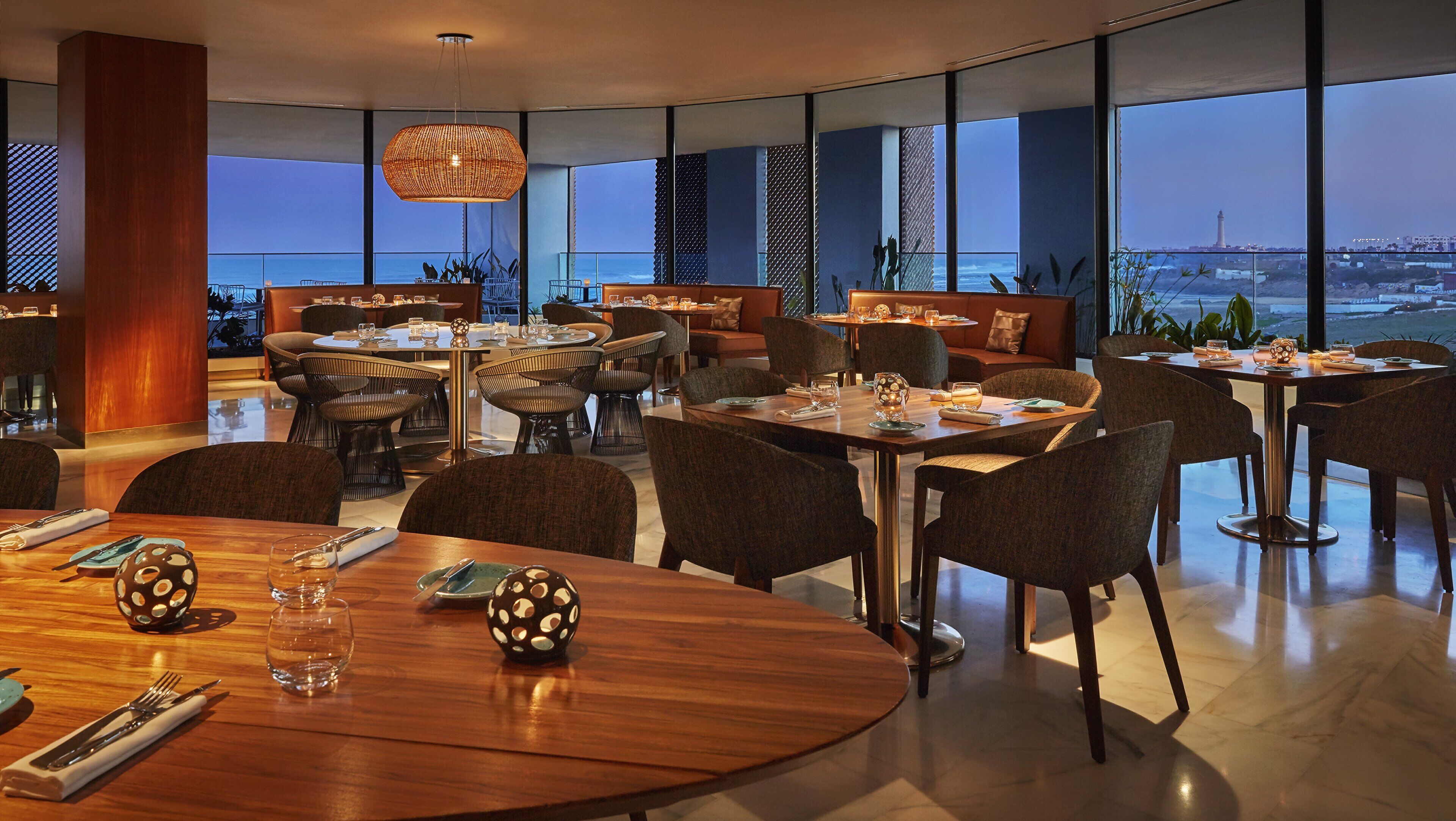 Un restaurant moderne avec des tables et des chaises rondes, de grandes fenêtres donnant sur l’océan et un luminaire suspendu.
