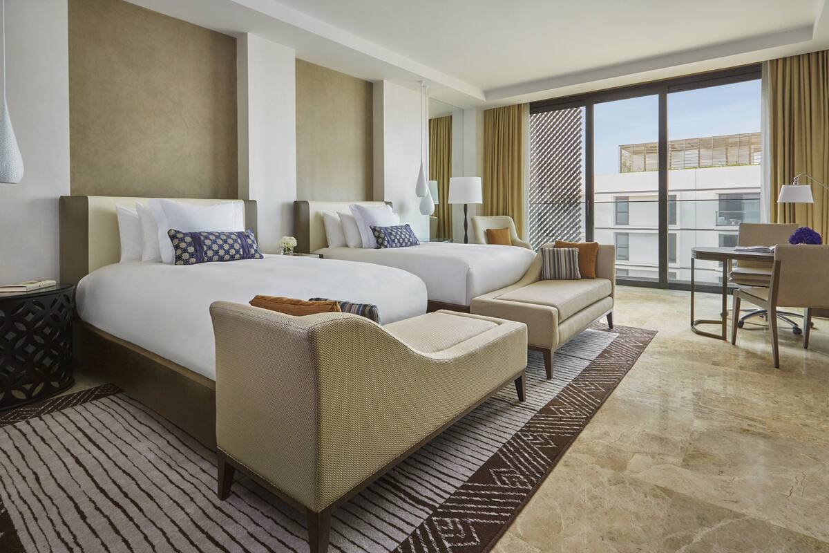 A hotel room with two beds, a sofa, a desk, and a large window with a view of buildings.