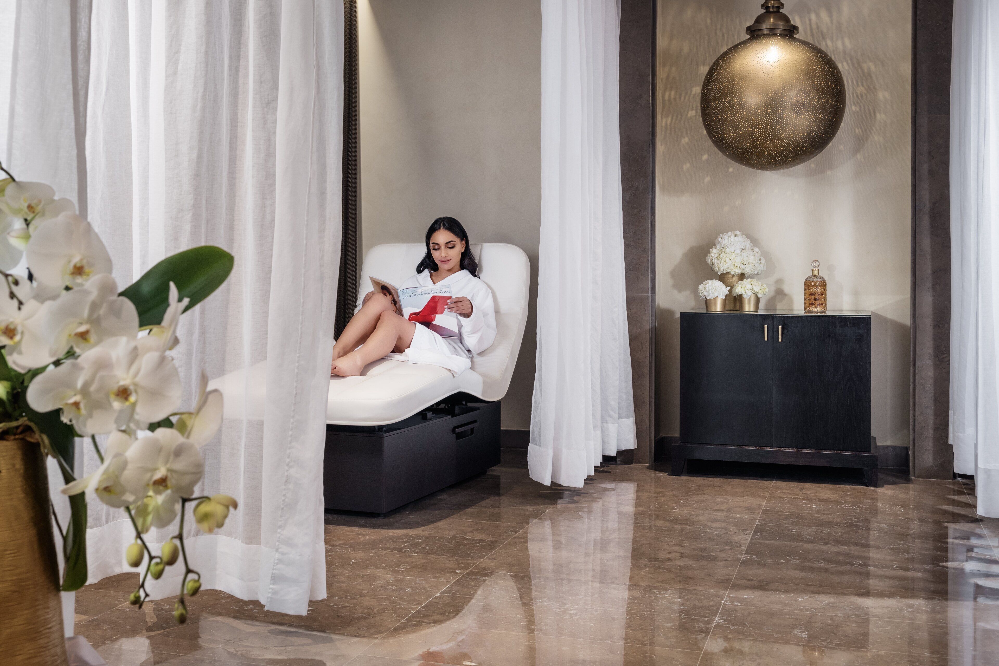 Ein Spa-Raum mit Massagesessel, einem schwarzen Schrank mit Dekoration und einer großen, goldfarbenen Pendelleuchte.