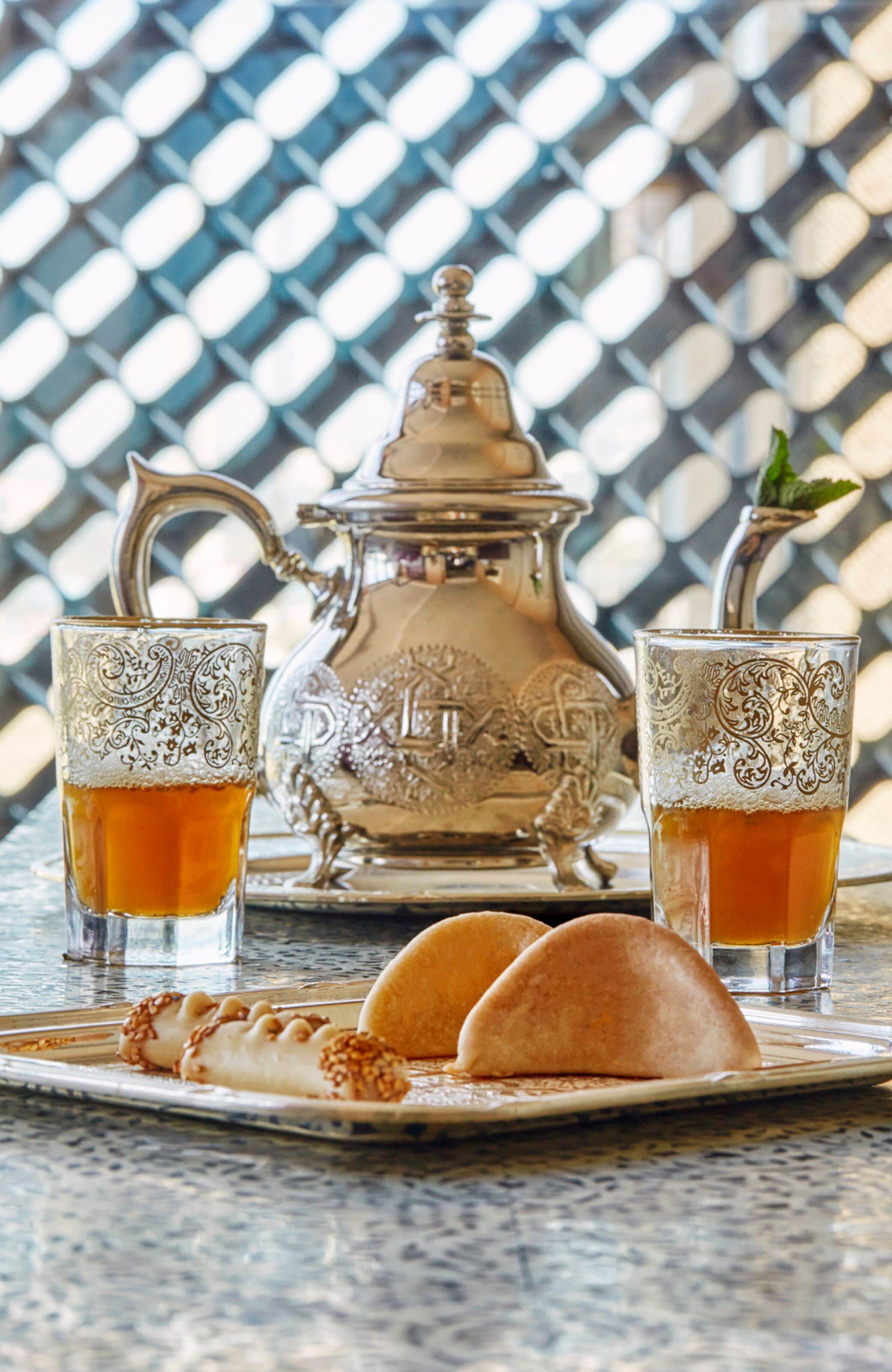 A tray with two glasses of tea, a silver teapot, and two pastries on a table with a patterned background.