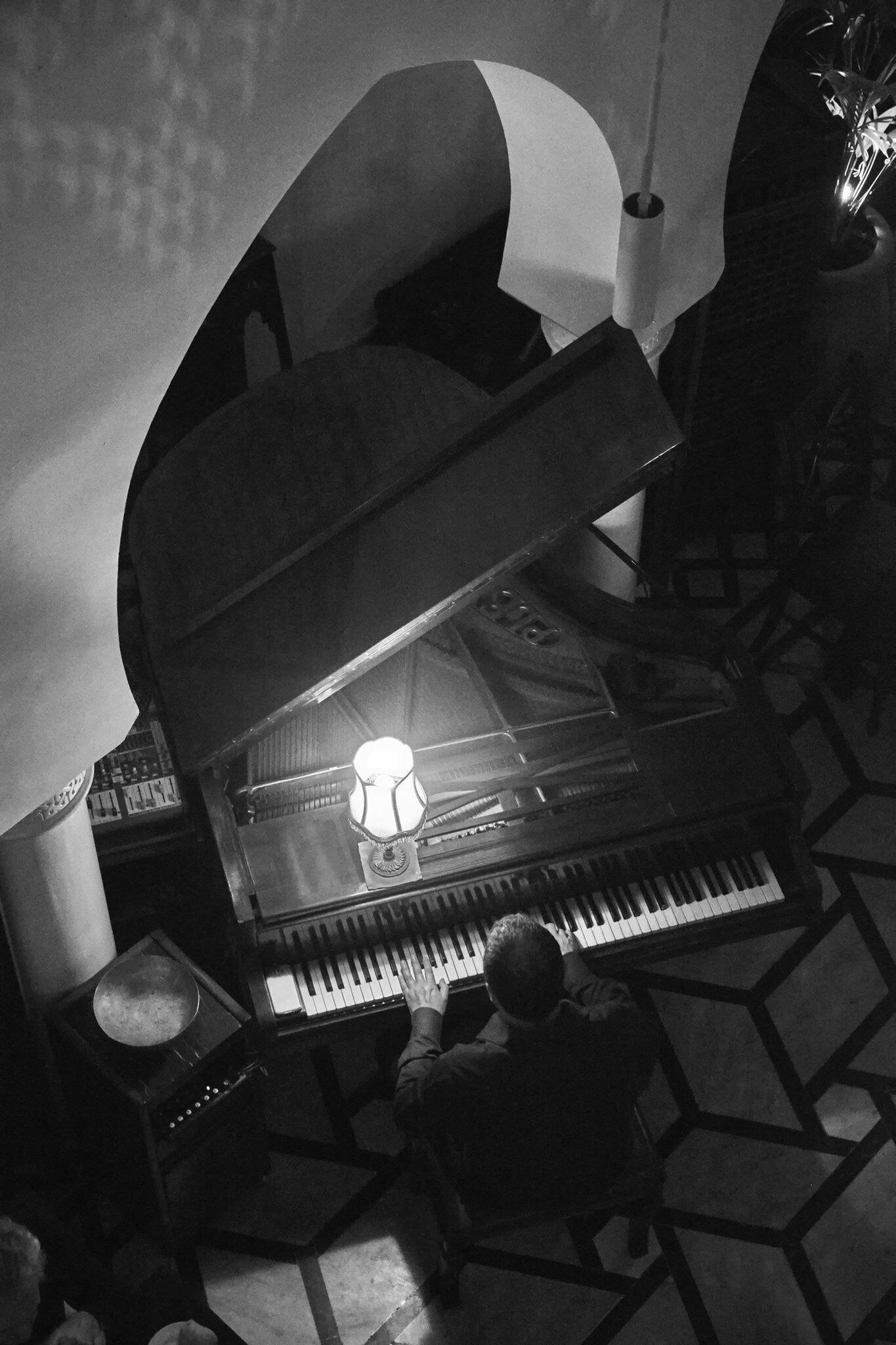 Een vleugelpiano in een kamer met een vloer met een geometrisch patroon, een kleine tafel met een lamp, en een persoon die piano speelt.