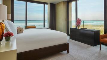 A hotel room with a large bed, a desk with a lamp, a chair, a small table with a vase, and a view of the beach.