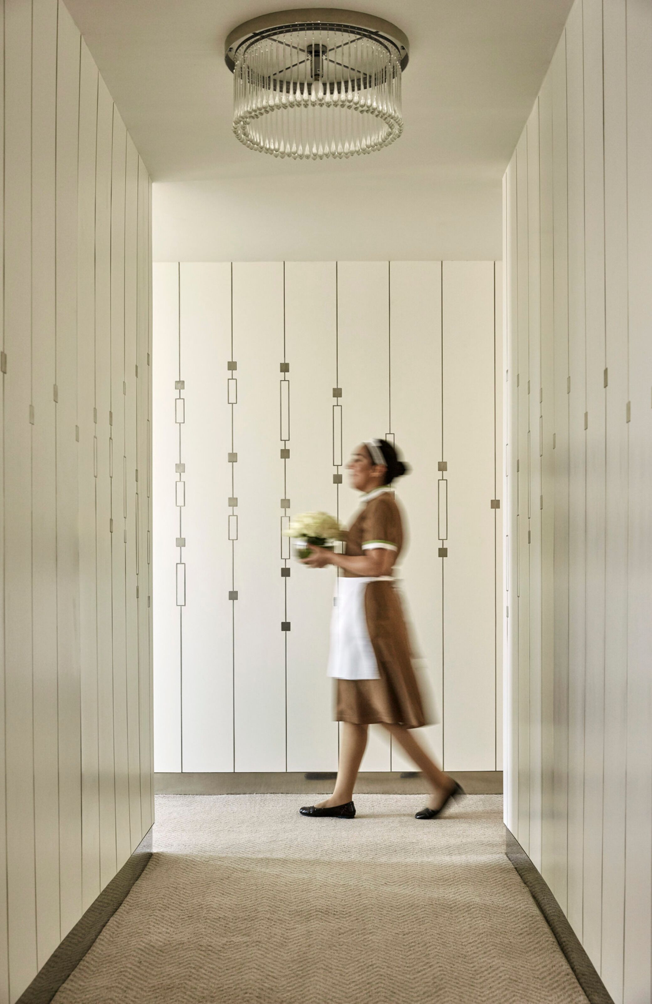 A hallway with a chandelier, lockers, and a person holding flowers.