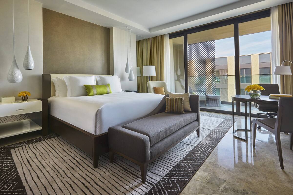A modern hotel room with a large bed, a sofa, a desk, and a view of the city through large windows.