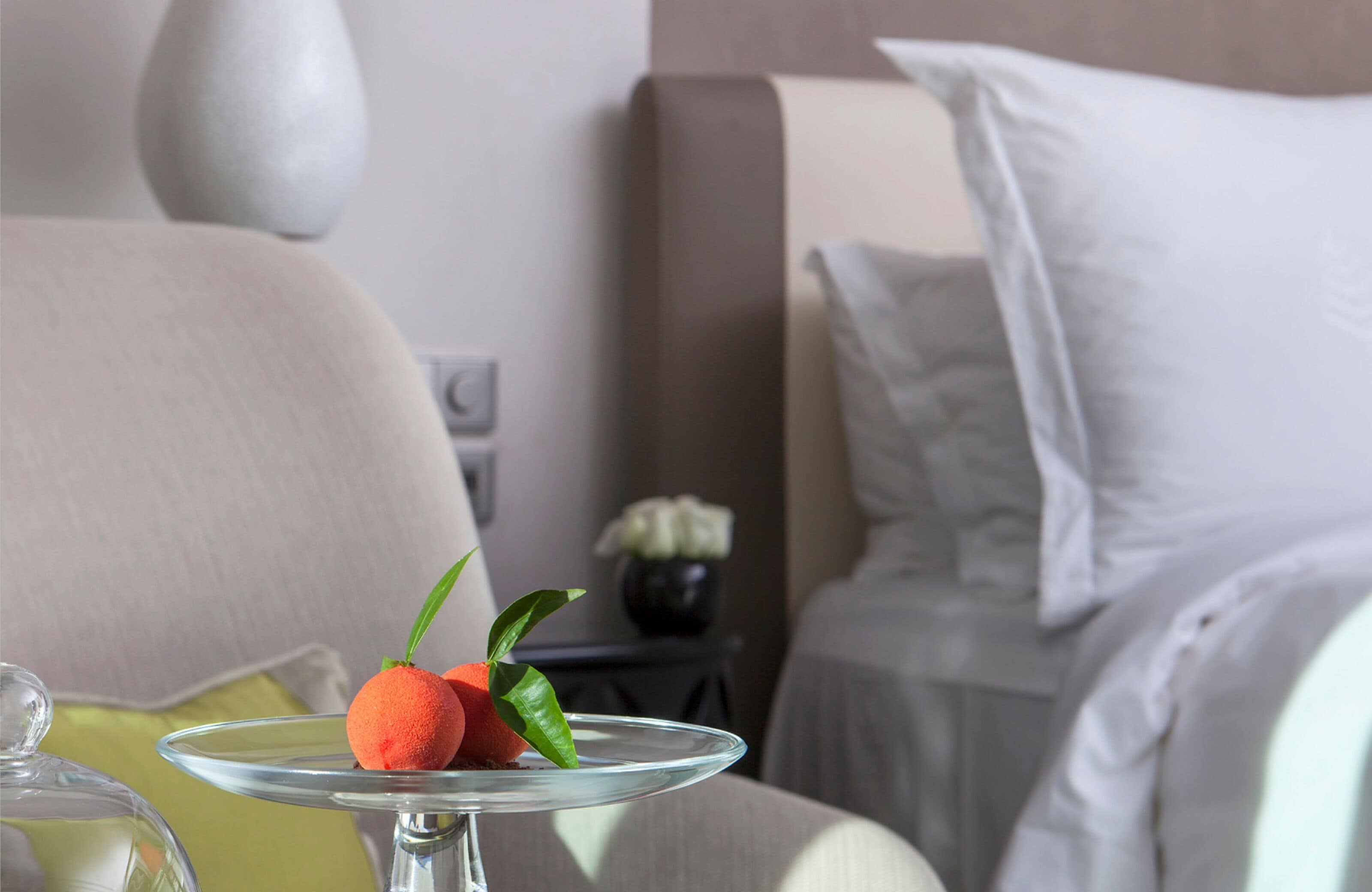 Habitación de hotel con una cama, una mesita de noche con un florero y una mesa de vidrio con decoración de frutas.