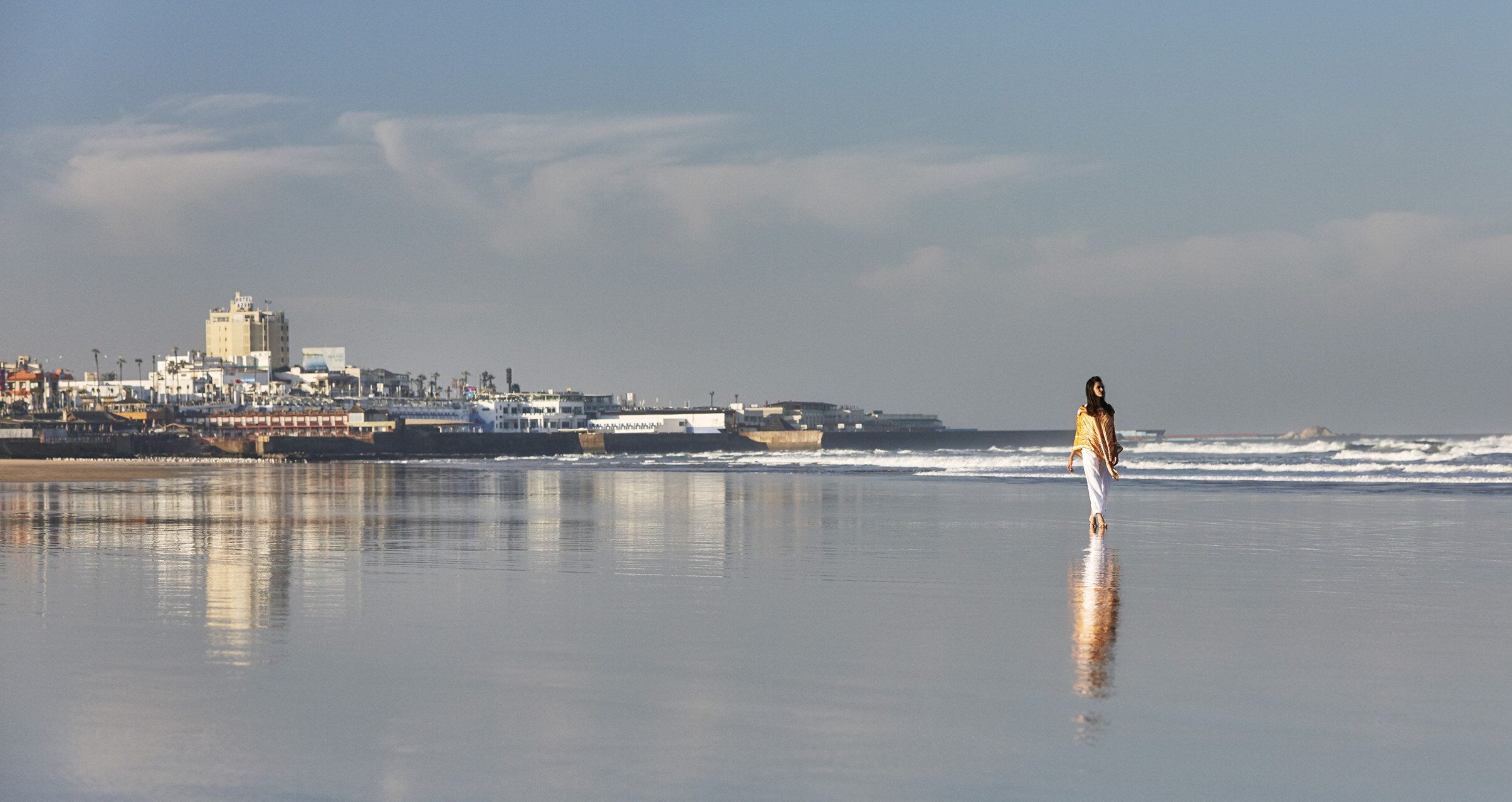 A beach with a person walking, a cityscape in the background, and a calm sea.