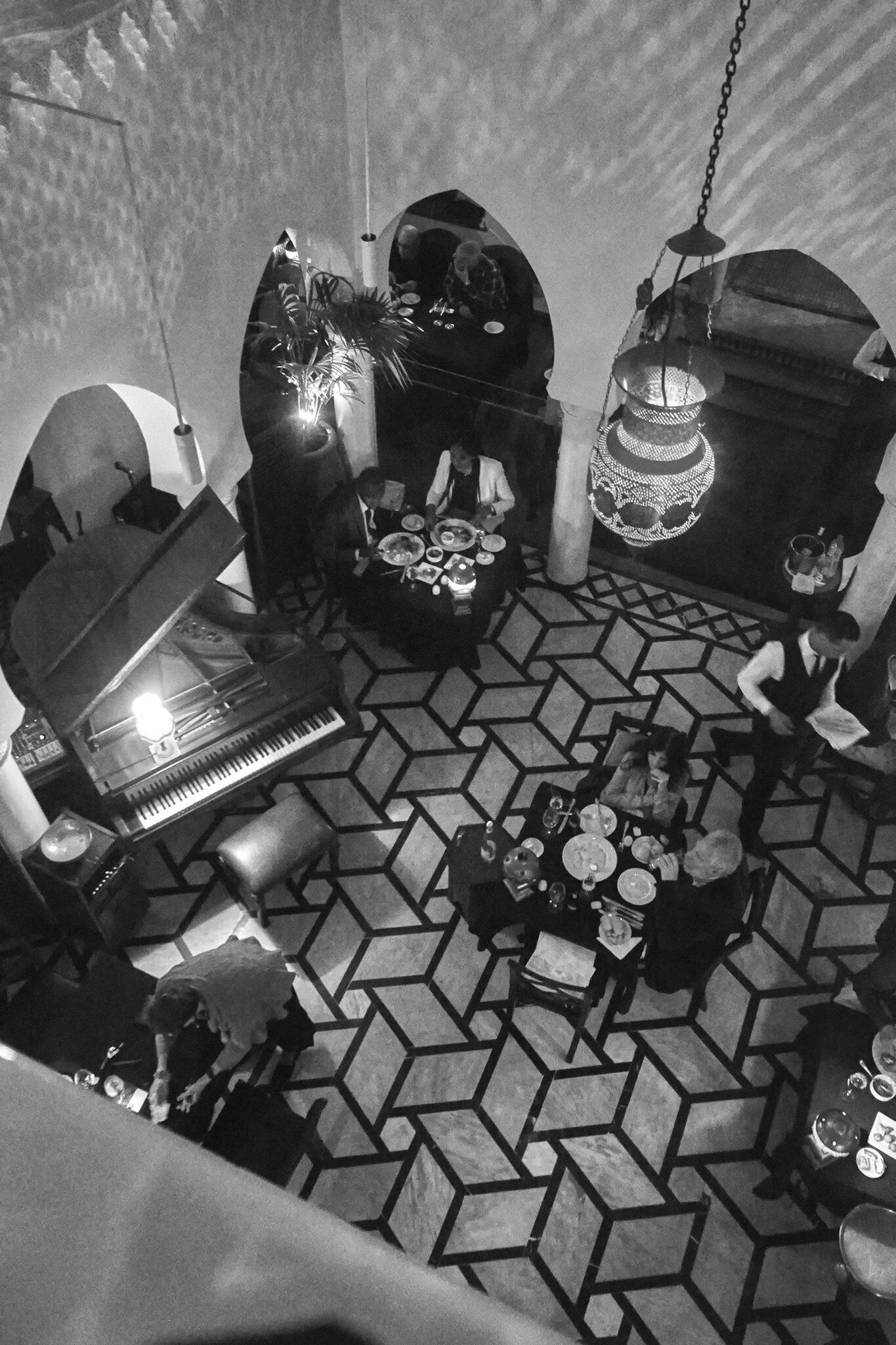 A room with a piano, a dining table with chairs, a chandelier, and a patterned floor.