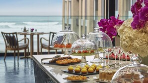 Una mesa de buffet con diversos postres y pasteles, una cúpula de vidrio que cubre algunos artículos, y vista al océano al fondo.