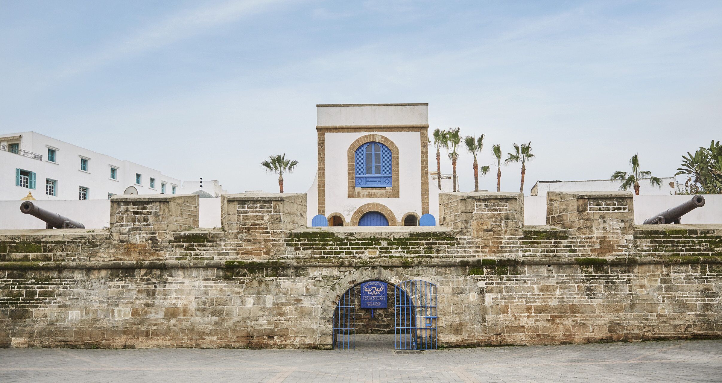 A historic fort with a central arched entrance, cannons on either side, and palm trees in the background.