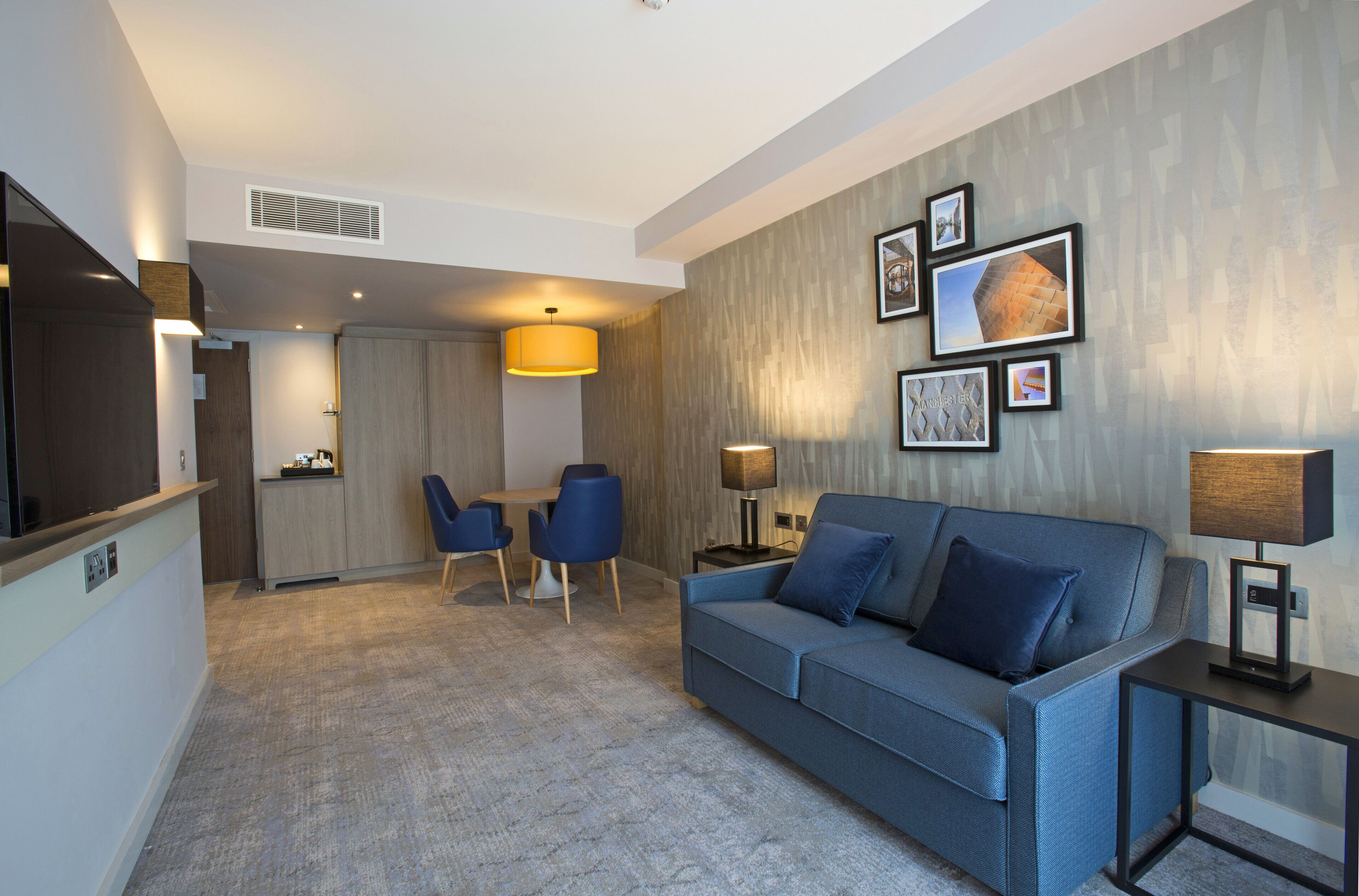 Una habitación de hotel moderna con sofá, una zona de comedor con sillas, un televisor y una lámpara.