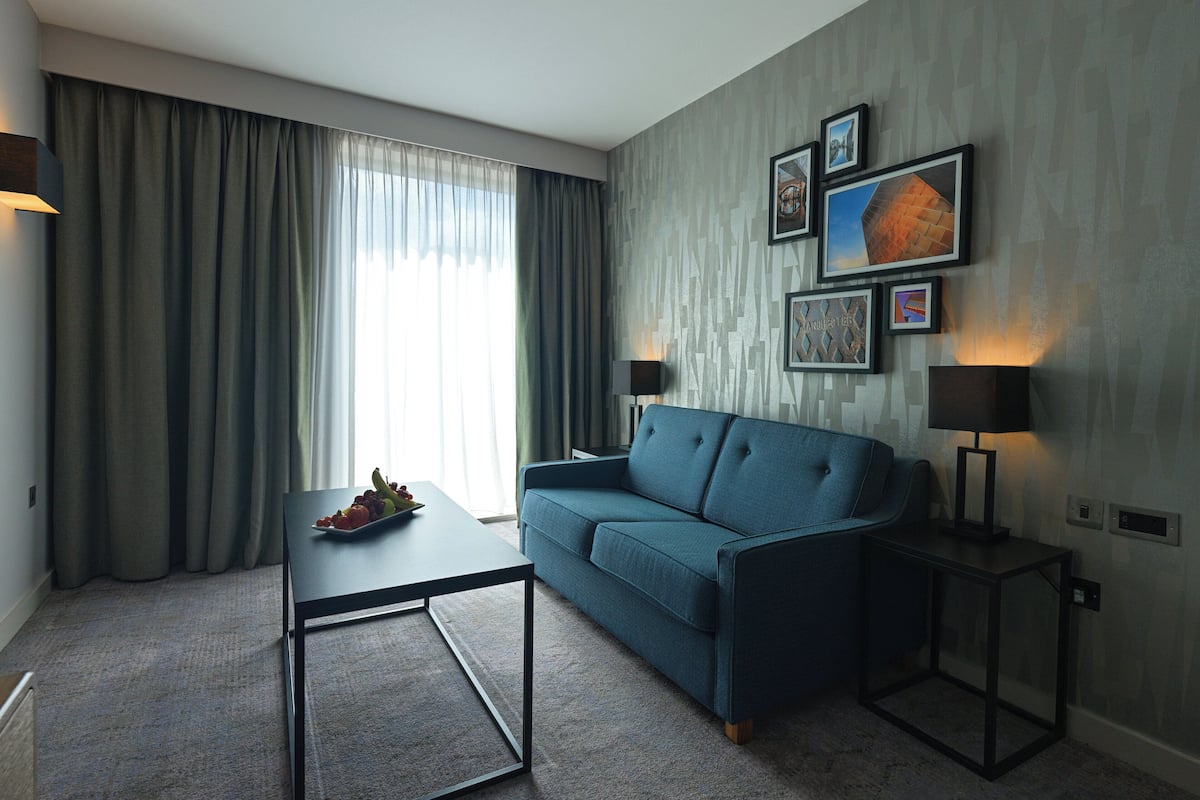 A modern living room with a blue sofa, a coffee table, and two side tables with lamps. There are curtains on the window, and a wall with framed pictures.