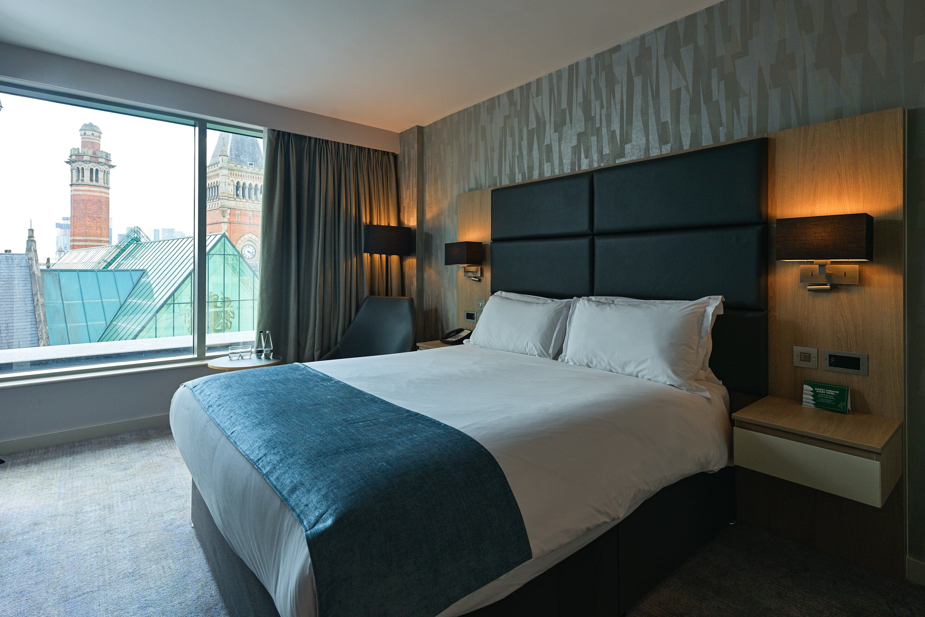 A hotel room with a large bed, two bedside tables with lamps, a view of a cityscape through a large window, and a patterned wall.