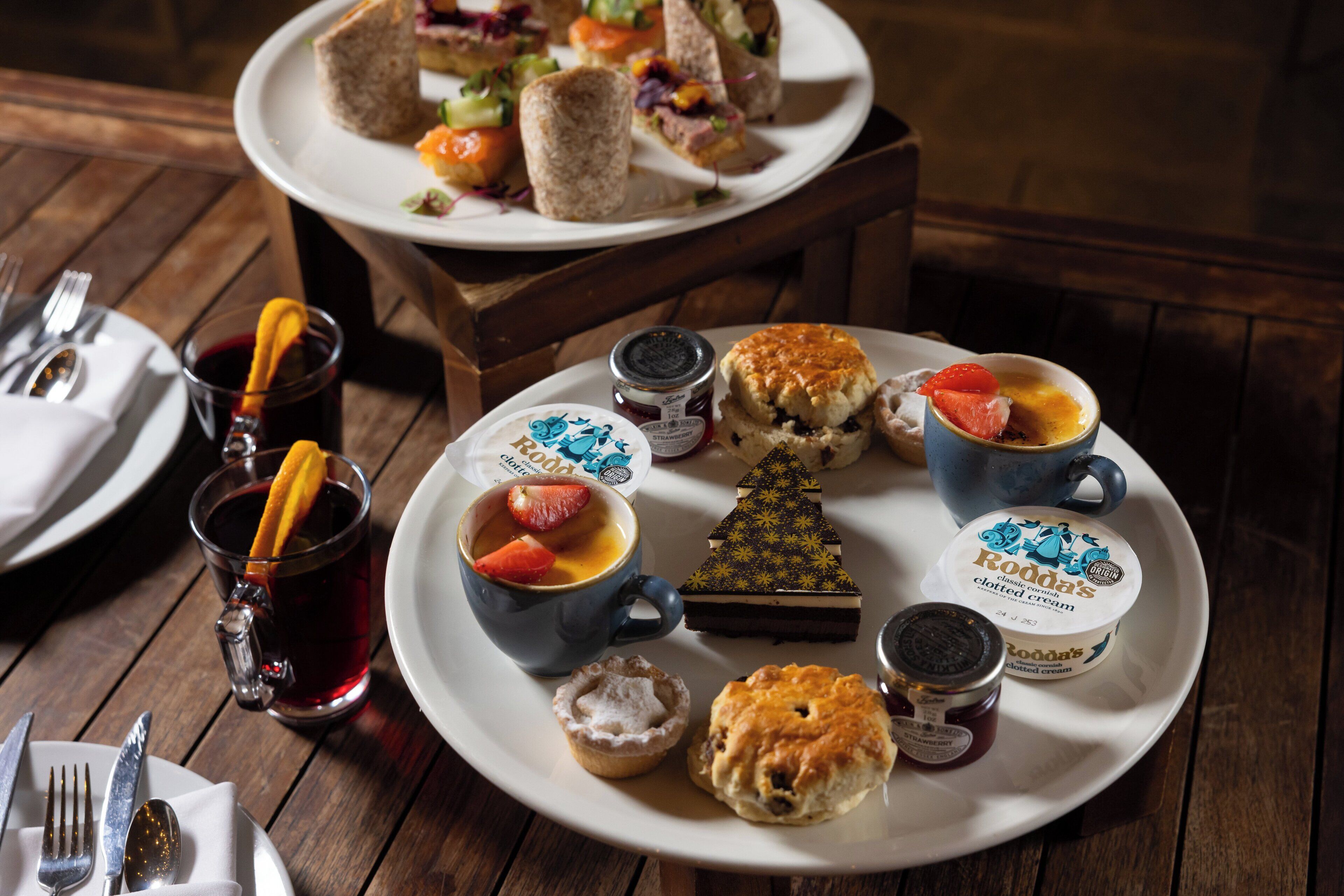 A dining table with a plate of sandwiches, a cup of tea, and a dessert tray.