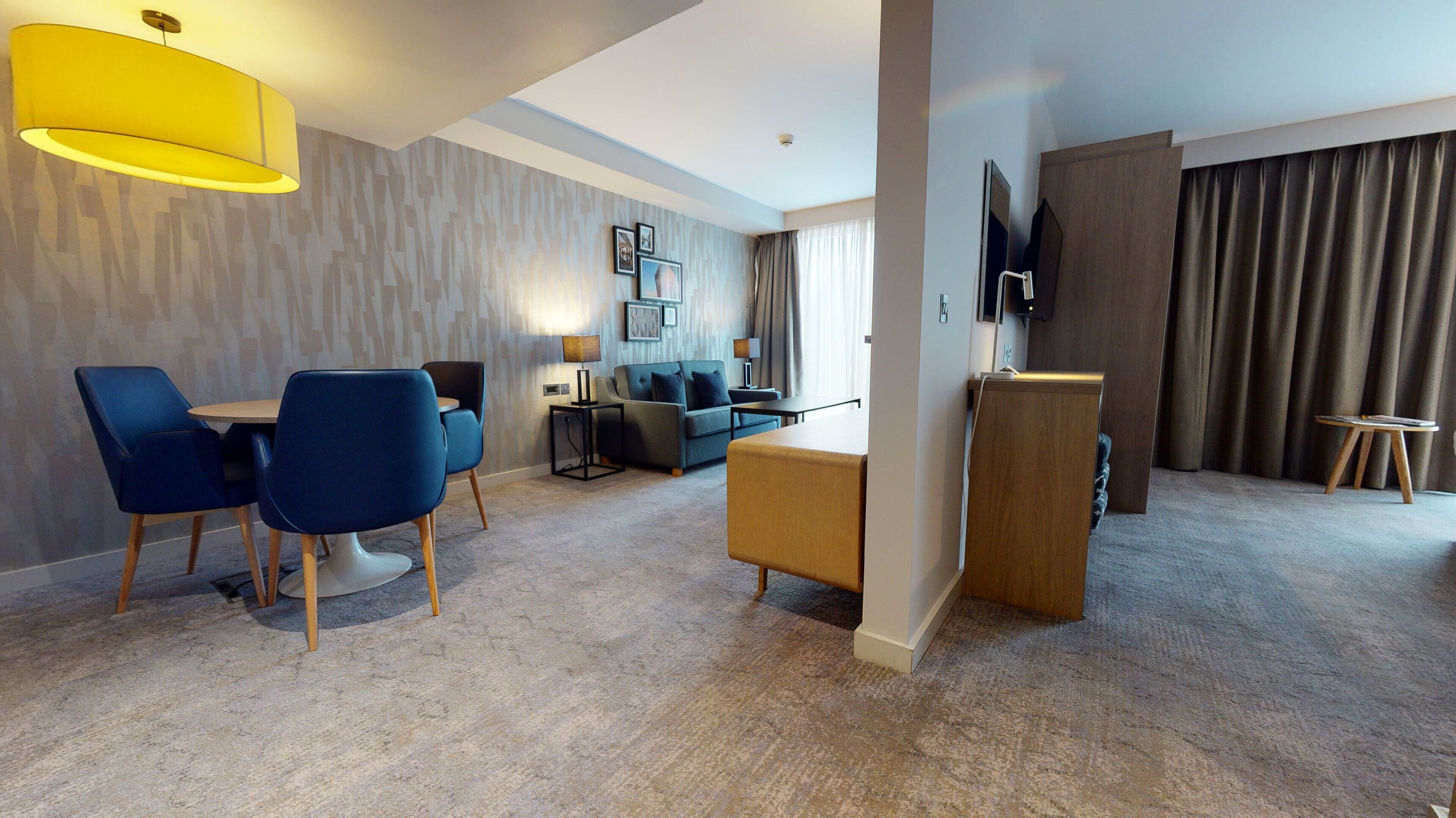 A modern hotel room with a dining area, a sofa, a TV, and a large window with curtains.