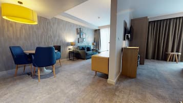 Una habitación de hotel moderna con zona de comedor, sofá, televisor y un amplio ventanal con cortinas.