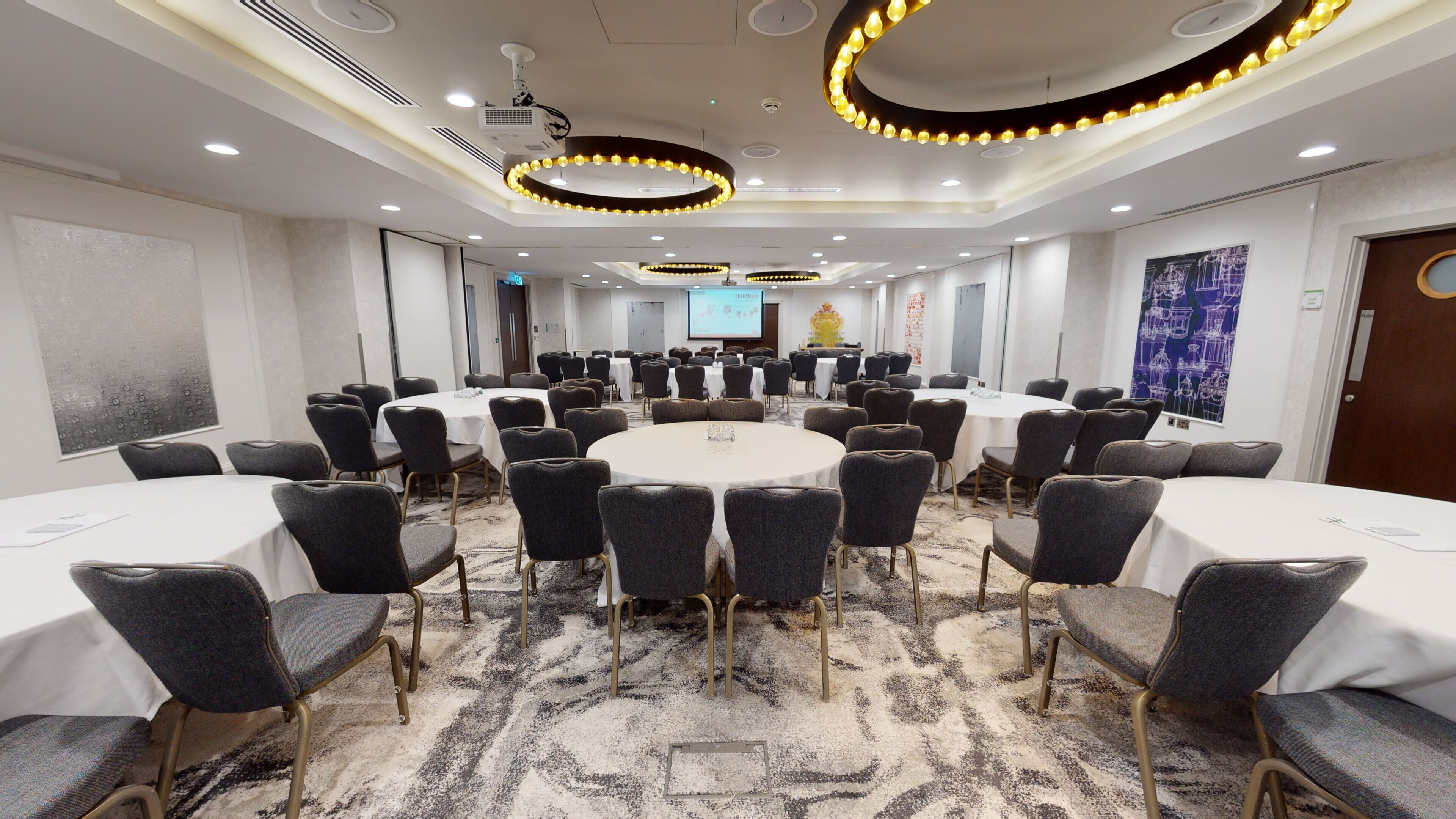 A conference room with round tables, chairs, a projector screen, and a large screen displaying a presentation.