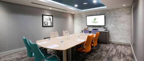 Una sala de conferencias con una larga mesa, sillas color verde azulado y naranja, un mapa en la pared y una pantalla mostrando 'Holiday Inn'.