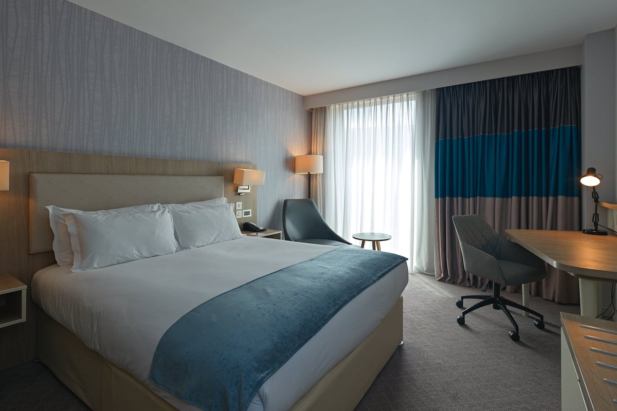 A hotel room with a large bed, two bedside tables, a desk with a chair, and a view of the outside through curtains.