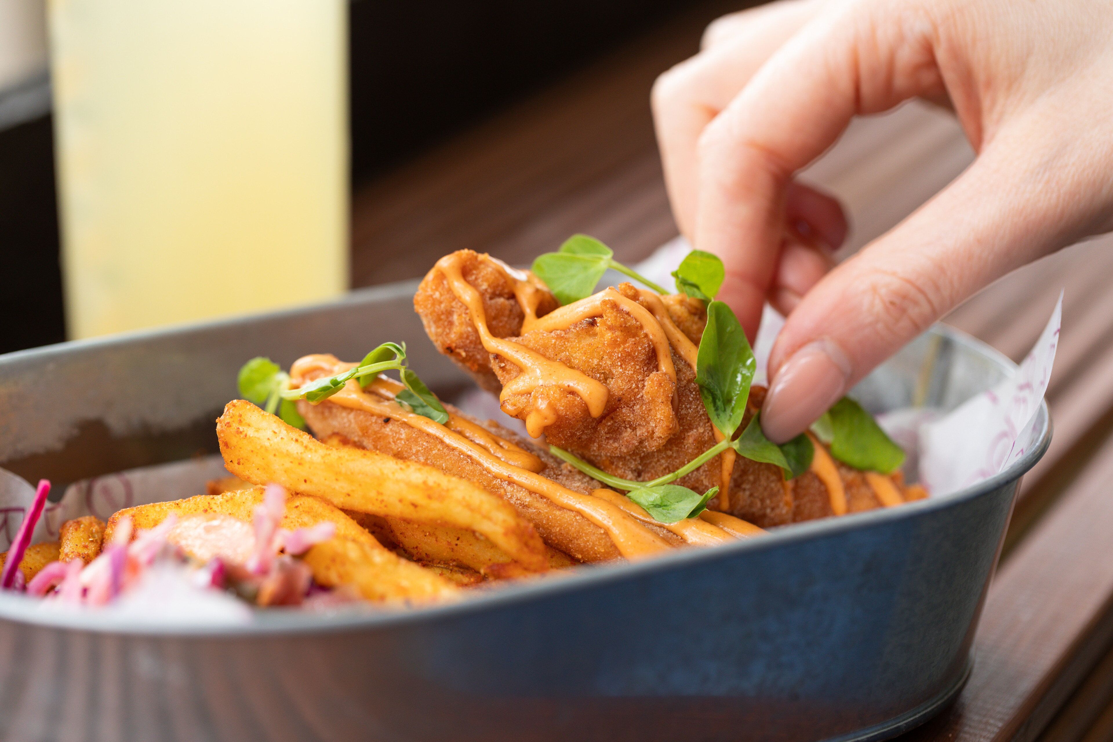 A hand holding a piece of fried food over a bowl containing fries and greens.