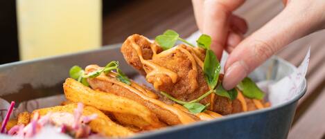 Una mano sostiene un bocado frito sobre un tazón con papas fritas y vegetales verdes.
