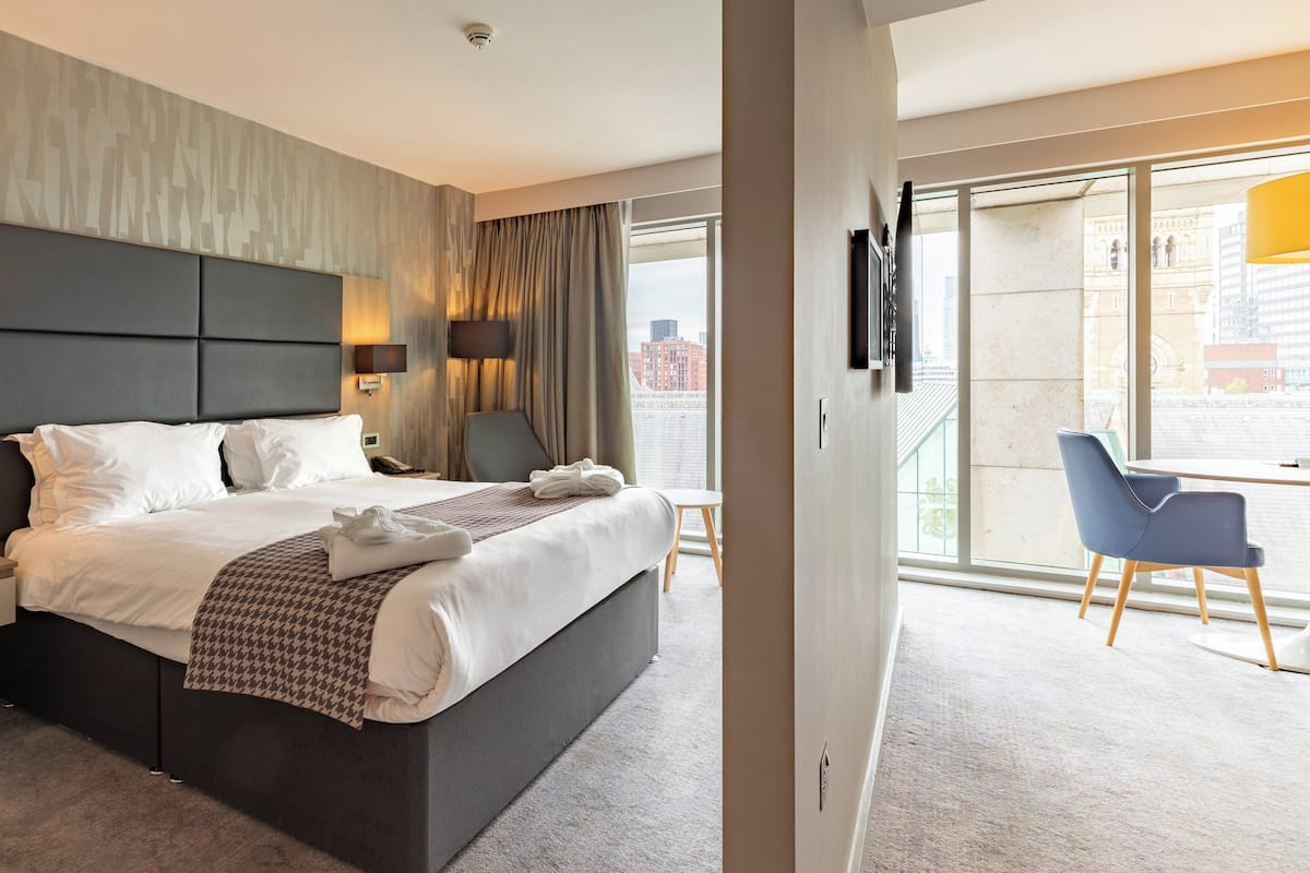 A modern hotel room with a large bed, bedside lamps, a flat-screen TV, a chair, and a view of the city through large windows.