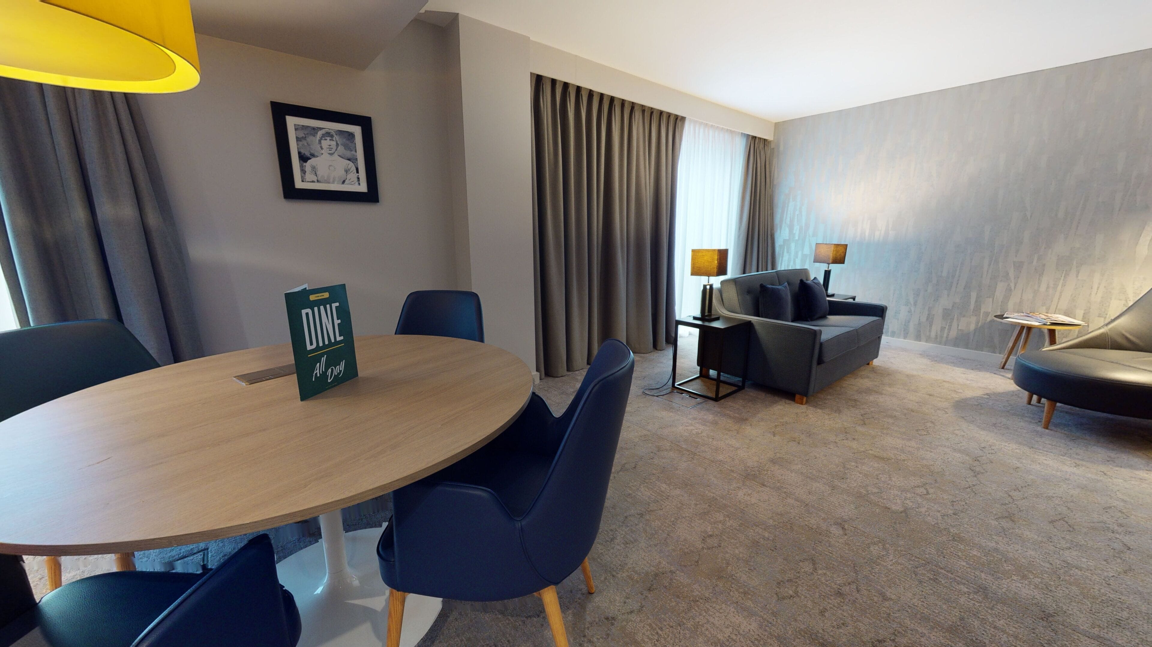 Een hotelkamer met een ronde houten tafel, twee blauwe stoelen, een bank, een klein bijzettafeltje met een lamp en een ingelijst schilderij aan de muur.