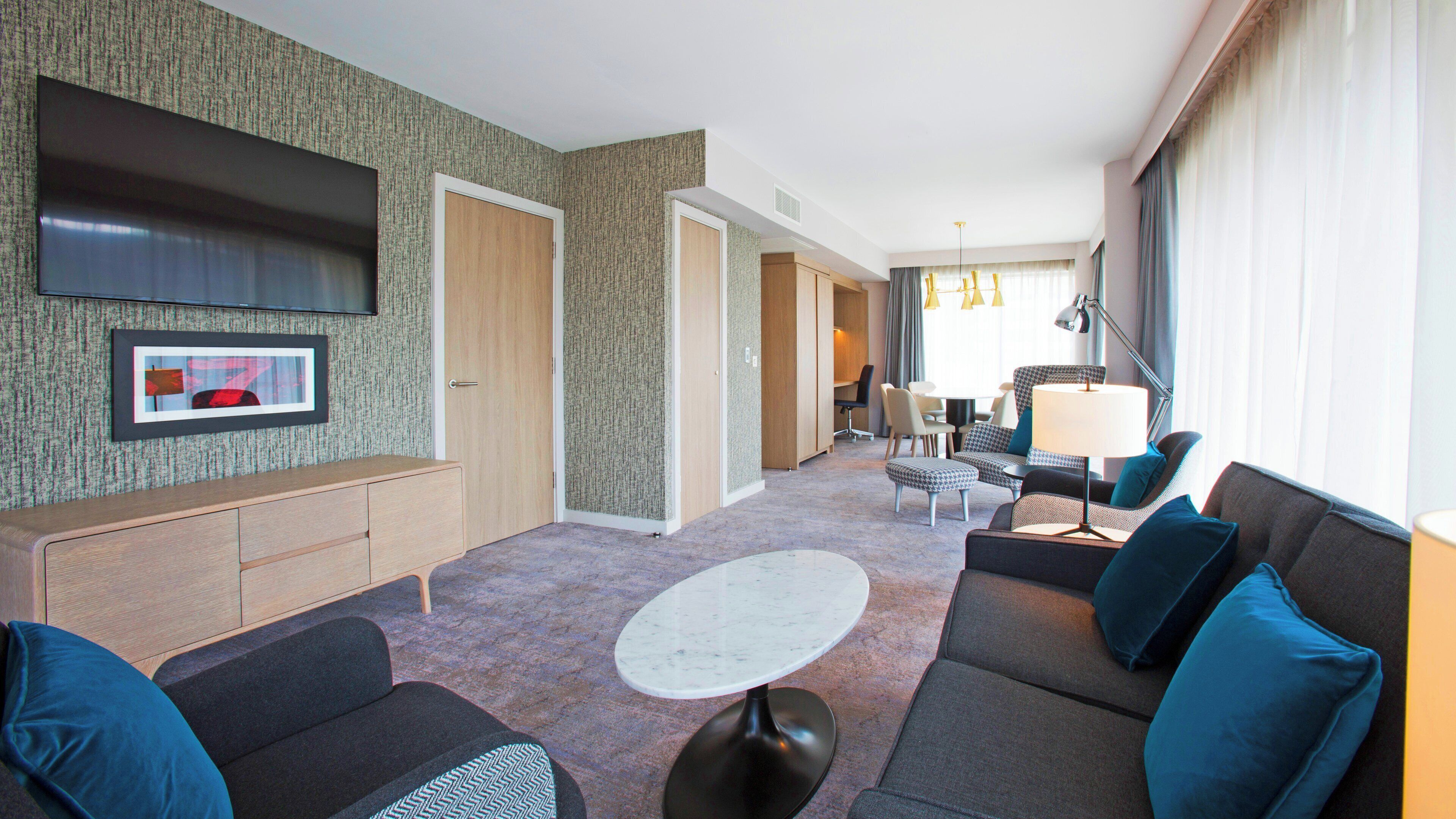 A hotel room with a seating area, a flat-screen TV mounted on the wall, a dining table with chairs, and a lamp.