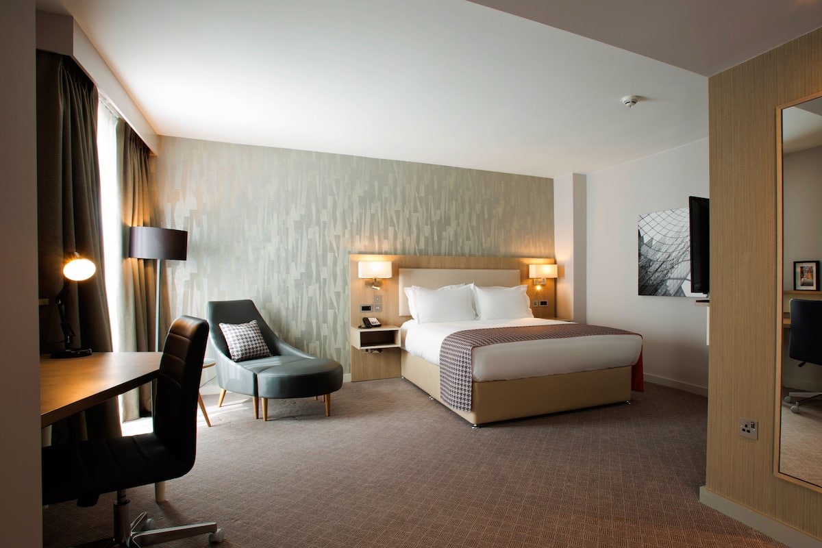 A hotel room with a bed, a desk with a chair, a bedside table with lamps, a large mirror, and a wall-mounted TV.