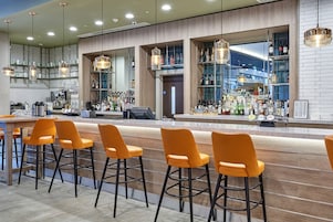 Un área de bar moderna con taburetes naranjas, una barra con una caja registradora y estantes llenos de botellas.