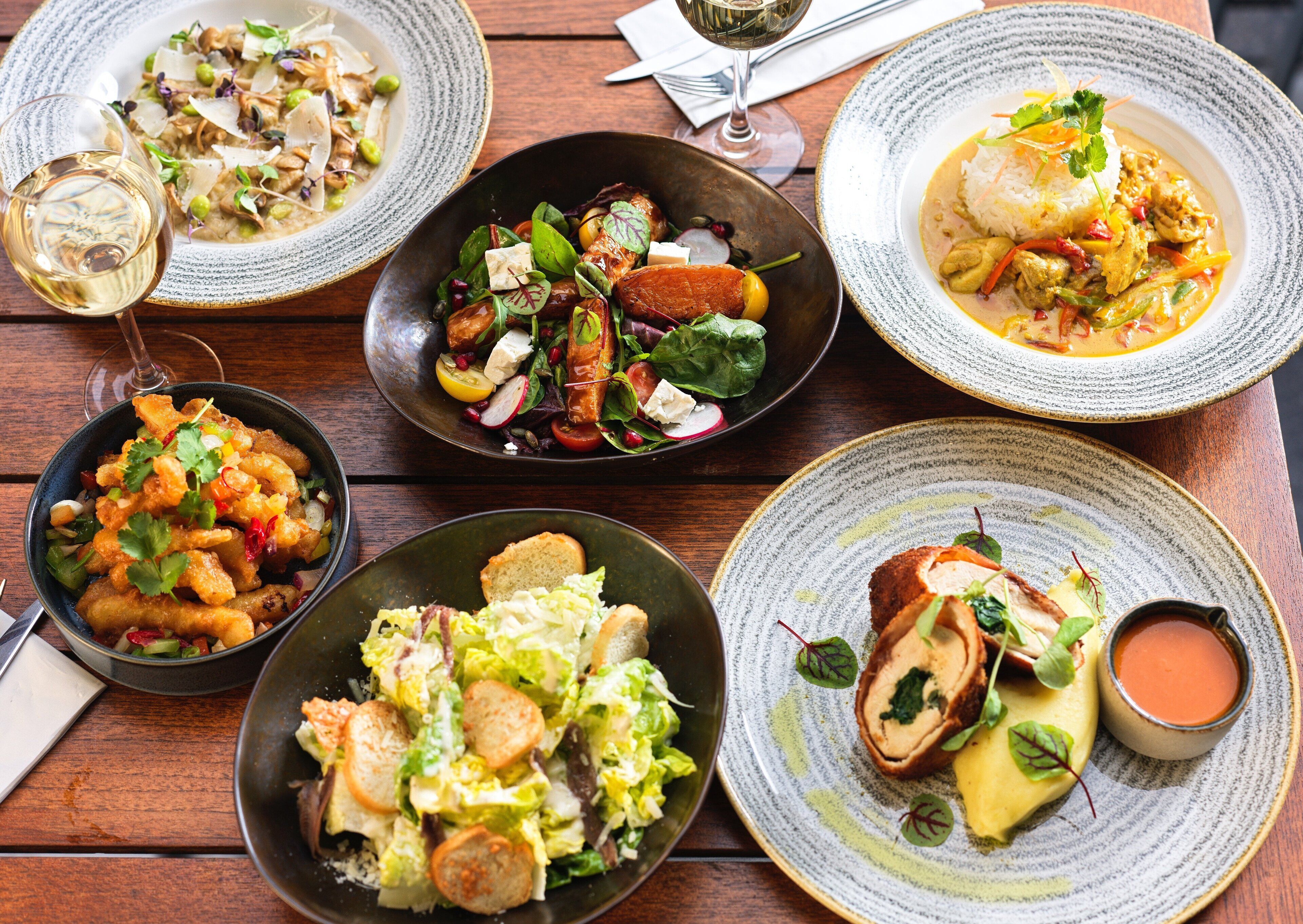 A variety of dishes on a wooden table, including salads, a soup, and a roll with sauce.