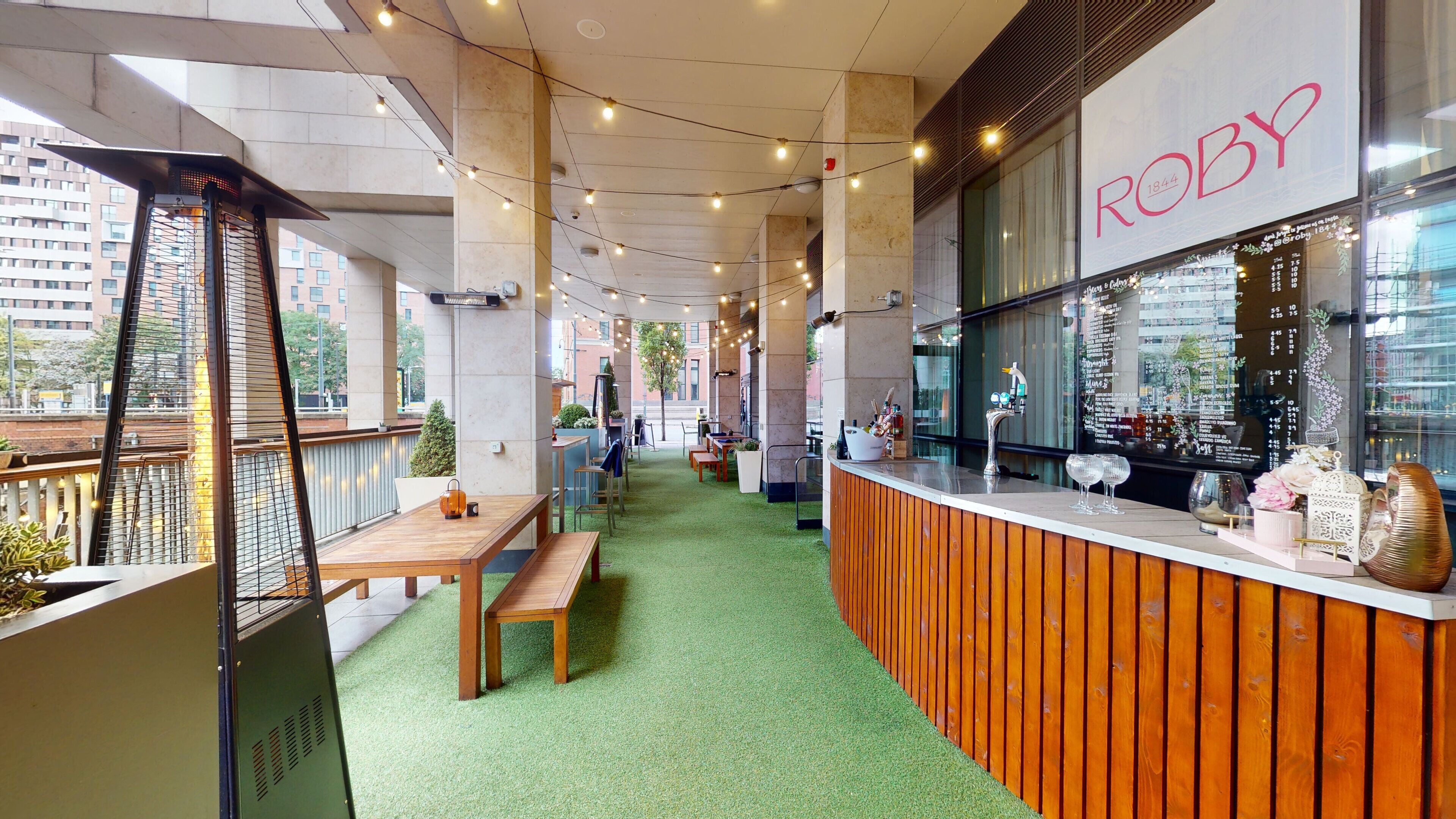 A restaurant with a wooden counter and menu board, green artificial grass flooring, and a view of the city.