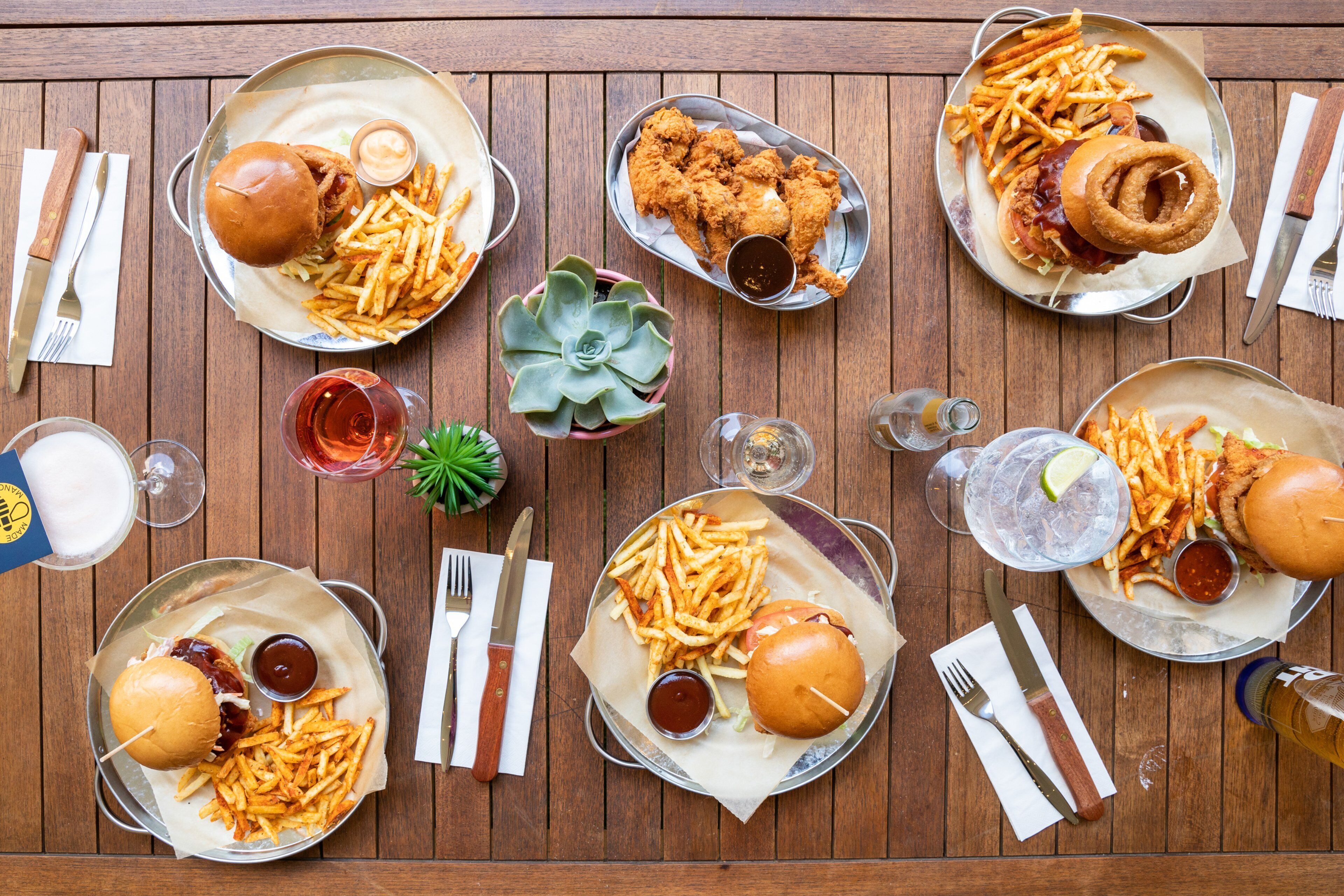 A wooden table with multiple plates of food including burgers, fries, and fried chicken, along with condiments and drinks.