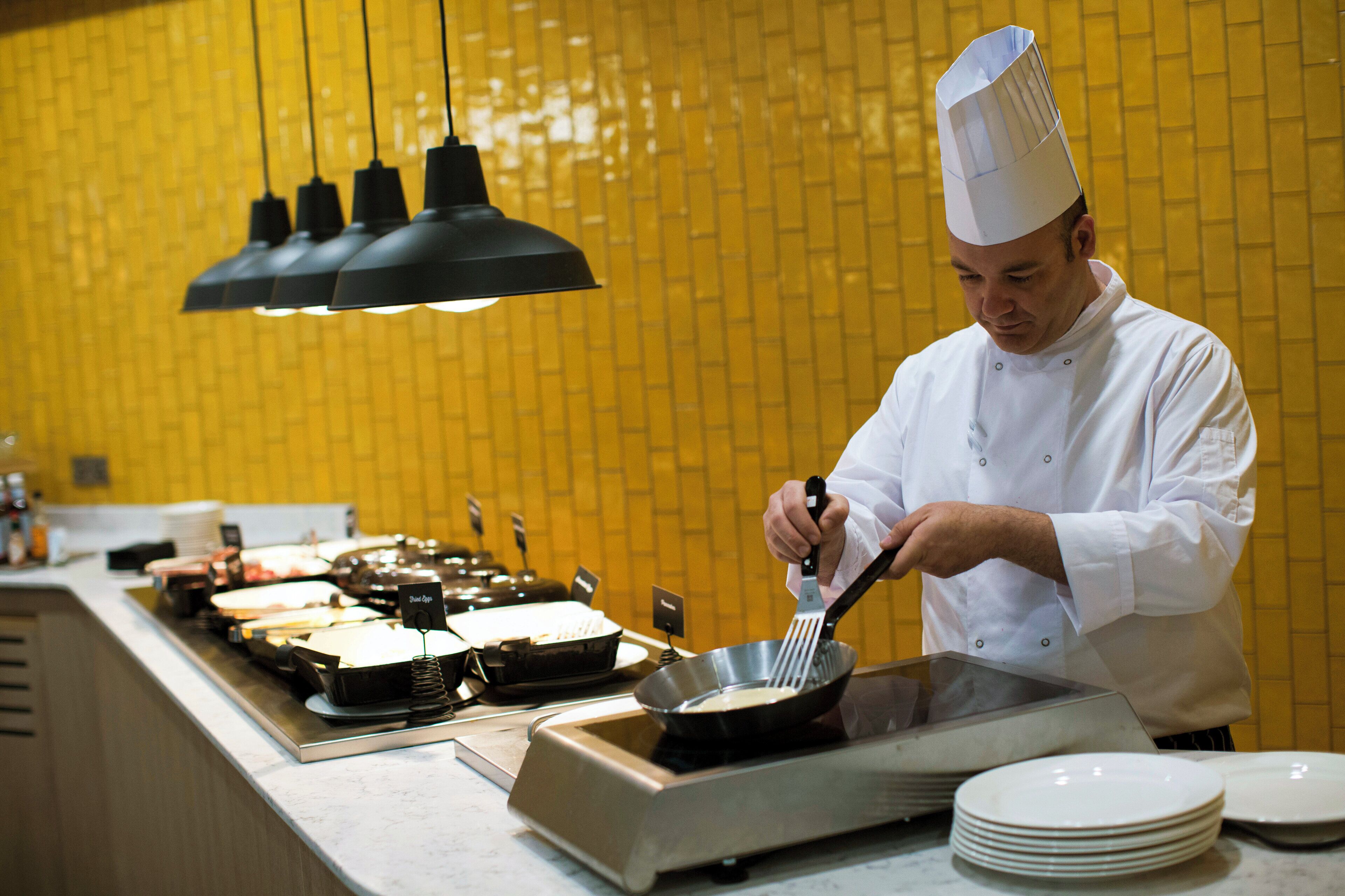 Un chef, vêtu d’un uniforme blanc, cuisine dans une cuisine aux murs recouverts de carreaux jaunes et éclairée par des suspensions noires.