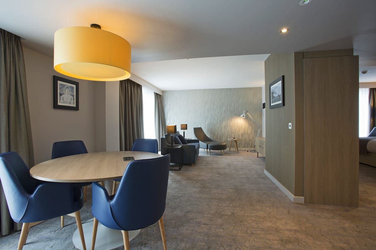 A modern hotel room with a round dining table, blue chairs, a sofa, and a lamp.