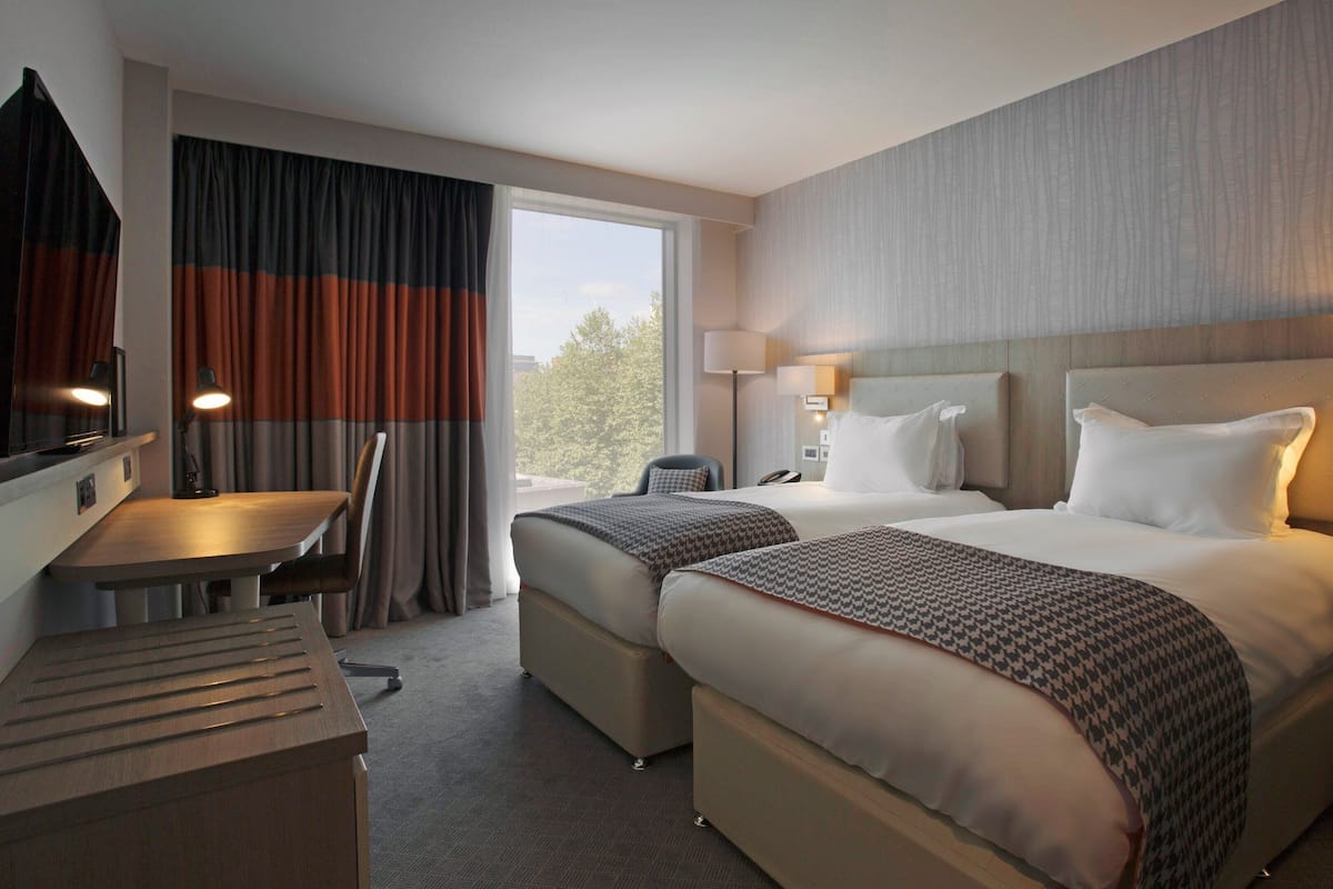 A hotel room with two beds, a desk, a chair, a TV, and a window with curtains.