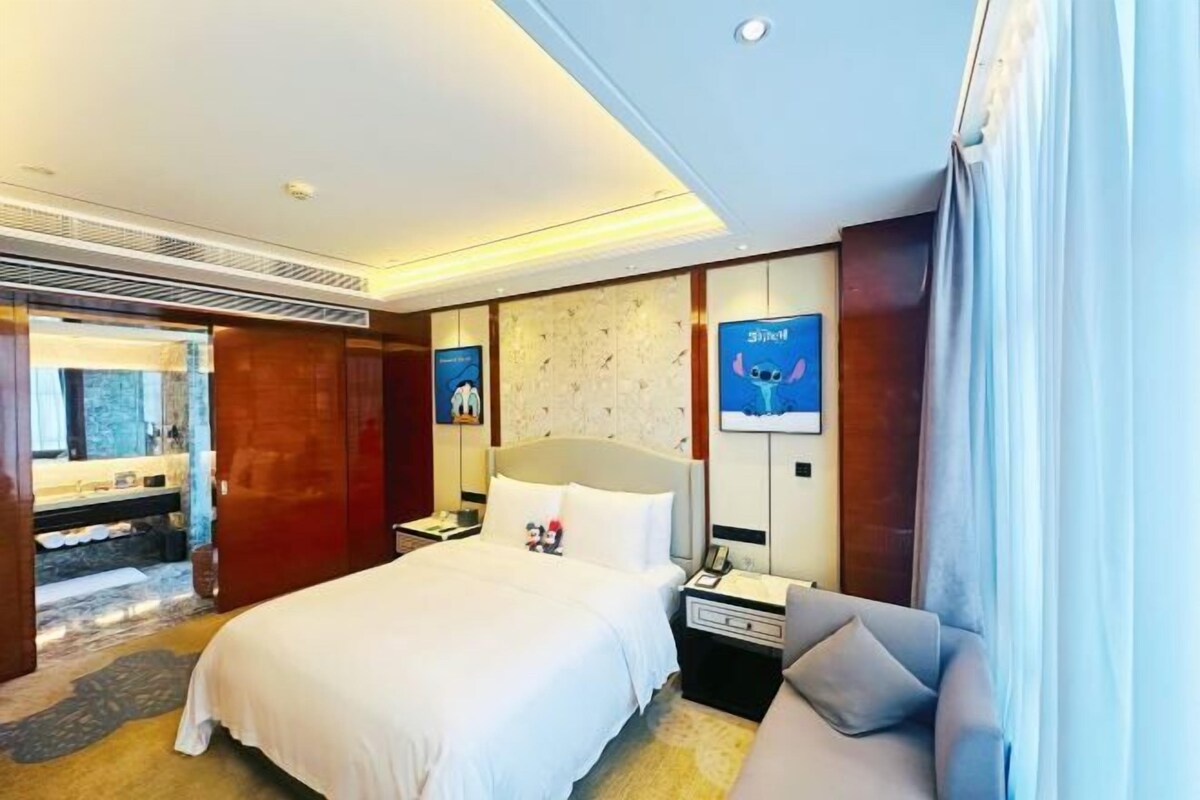 A hotel room with a bed, a nightstand, a sofa, a bathroom with a mirror, and a wall with framed pictures.