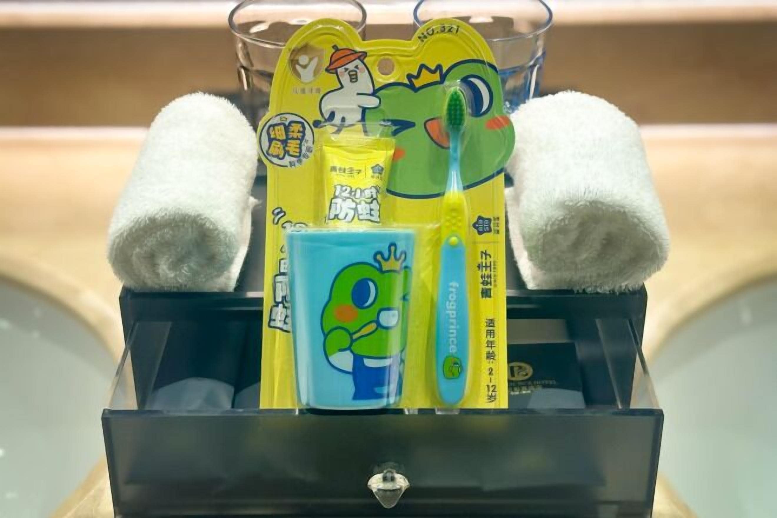 A drawer containing a toothbrush, toothpaste, a small blue cup with a frog design, and two white towels.
