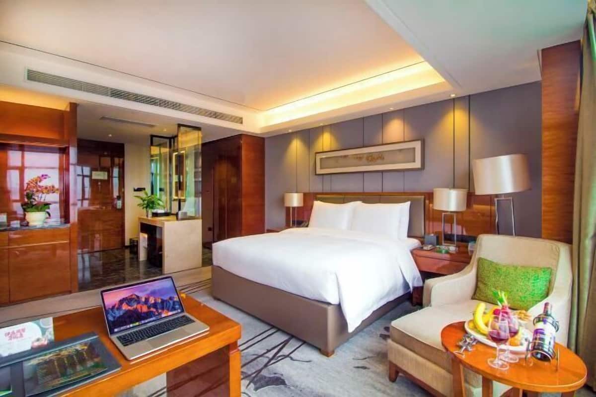 A hotel room with a large bed, a desk with a laptop, a chair, and a small table with a bottle and glasses.