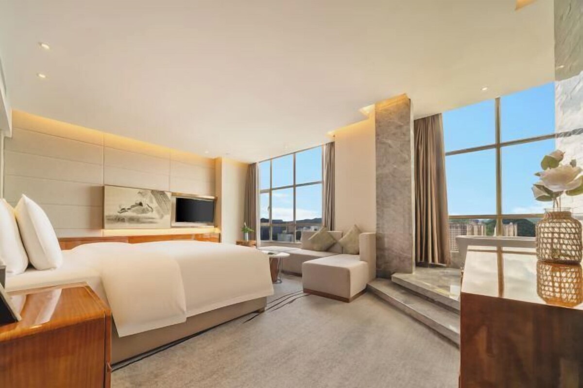 A modern hotel room with a large bed, a sofa, a TV, and a large window with a view.