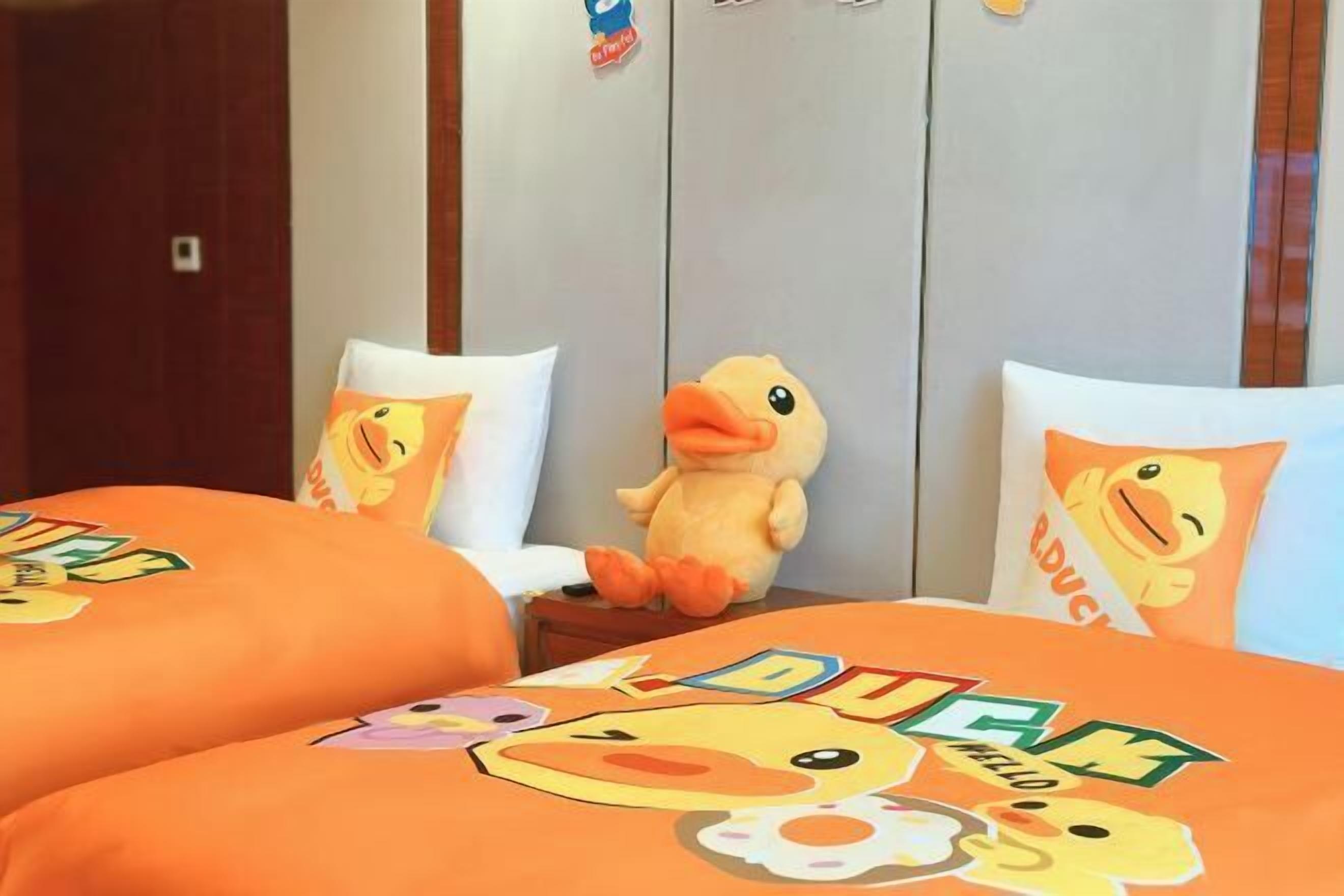 Une chambre d’hôtel avec deux lits, chacun agrémenté d’une literie orange ornée de motifs de canards. Une peluche en forme de canard est posée sur la table de chevet.