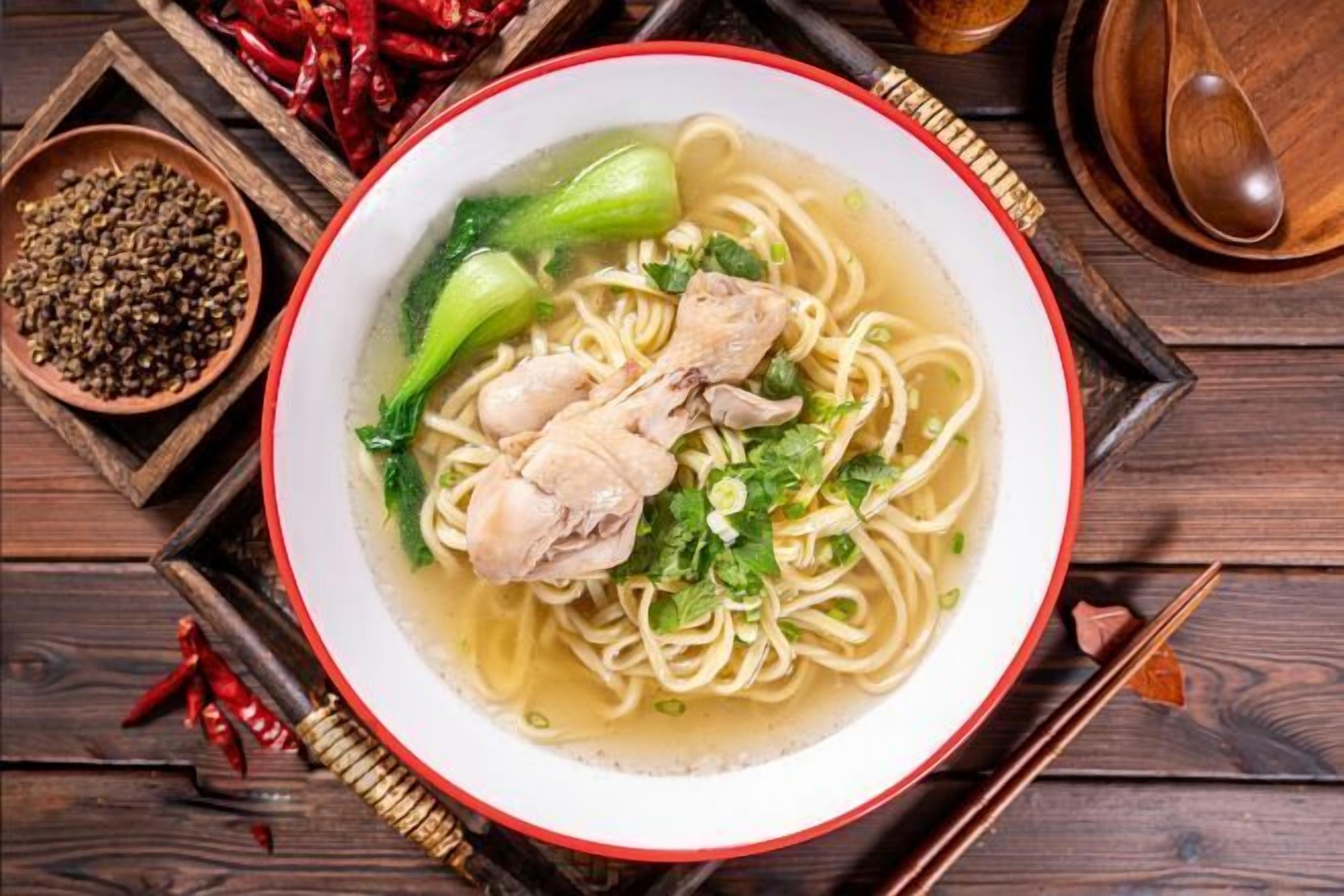 Un tazón de sopa de fideos con pollo y bok choy, rodeado de tazones de madera y chiles rojos.