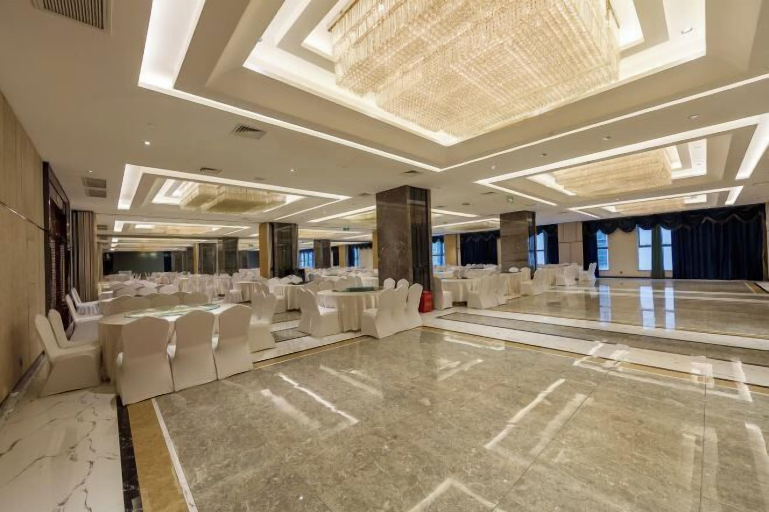 A spacious banquet hall with a polished marble floor, white round tables, and chairs, and a large chandelier.