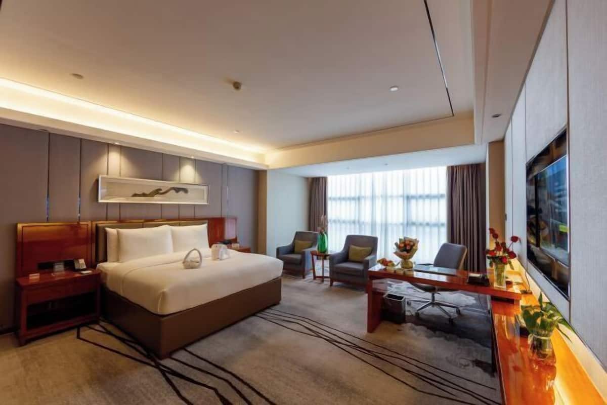 A hotel room with a large bed, a desk, two chairs, a TV, and a window with curtains.