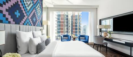 A modern hotel room with a large bed, a desk, two chairs, a TV, and a view of tall buildings.