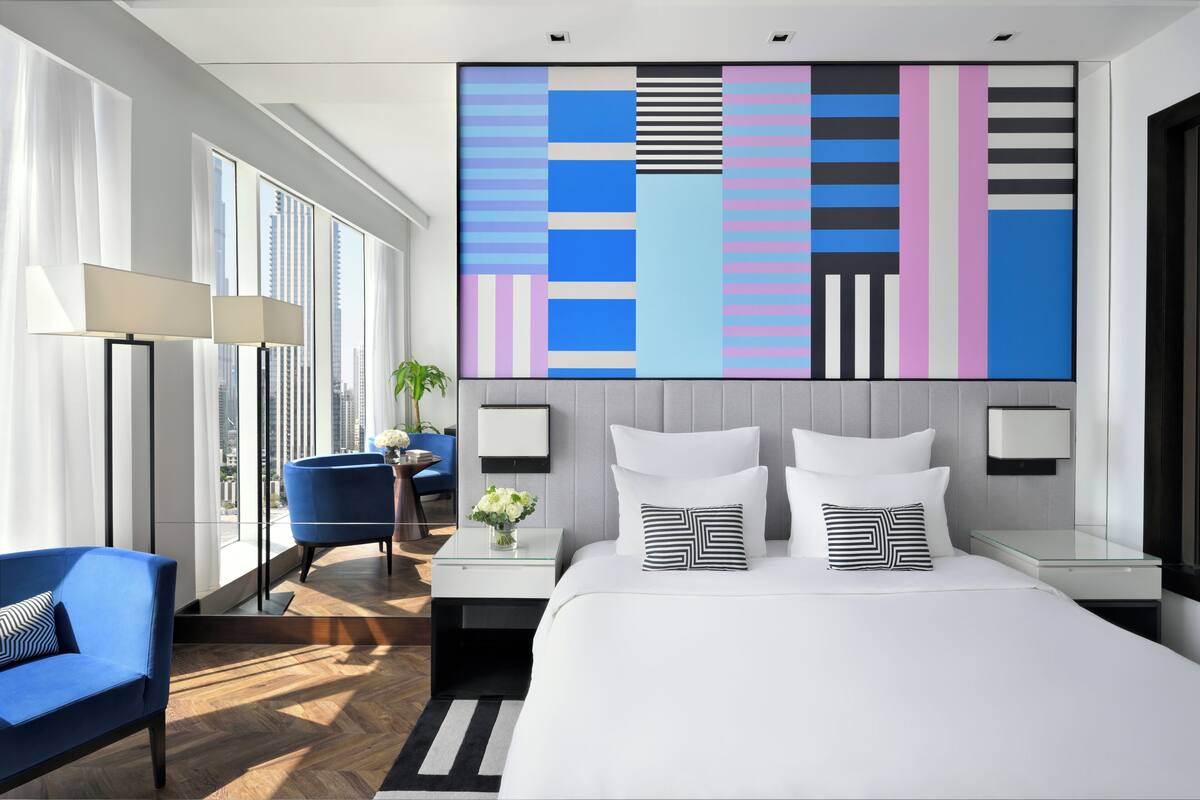 A modern hotel room with a large bed, a blue armchair, a glass coffee table, and a city view.