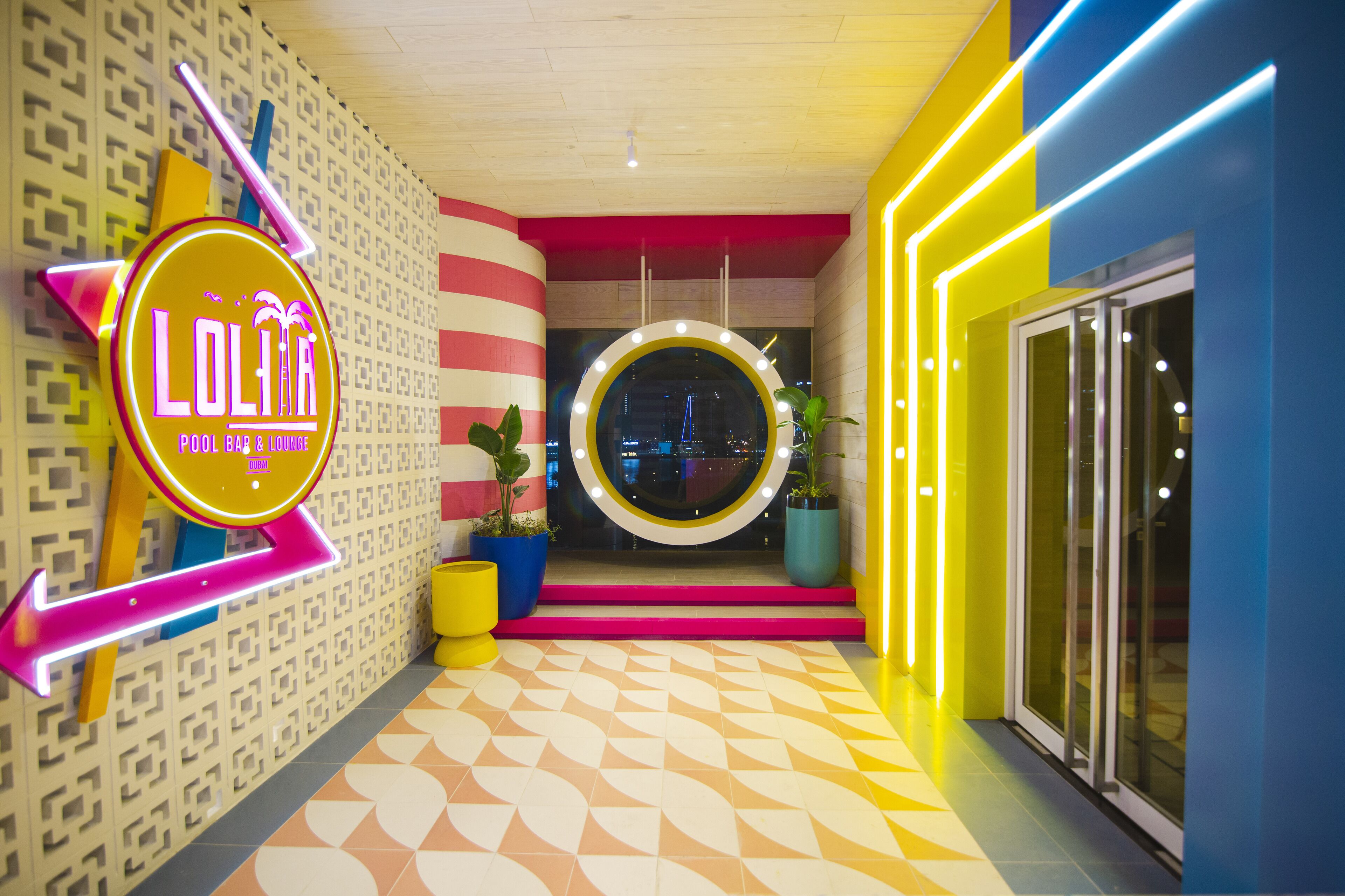 Um corredor bem iluminado com um letreiro de neon para 'LOLIA' e um espelho circular na parede.