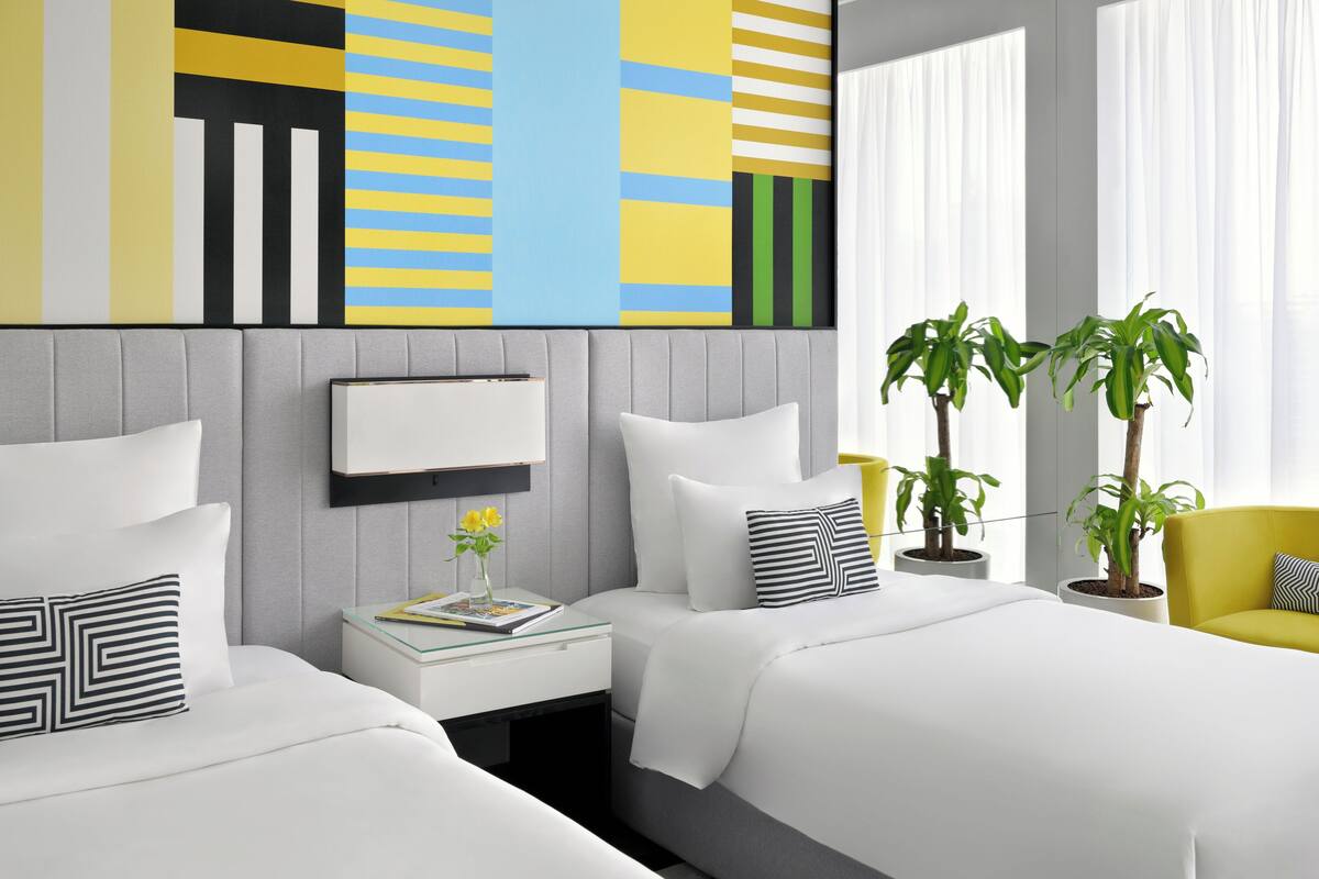 A modern hotel room with two beds, a bedside table with a book and vase, a large abstract painting, and two potted plants.